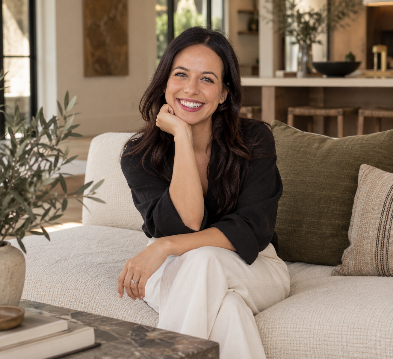 Woman with long dark hair sitting on a beige couch, smiling, in a bright living room with plants and wooden accents.