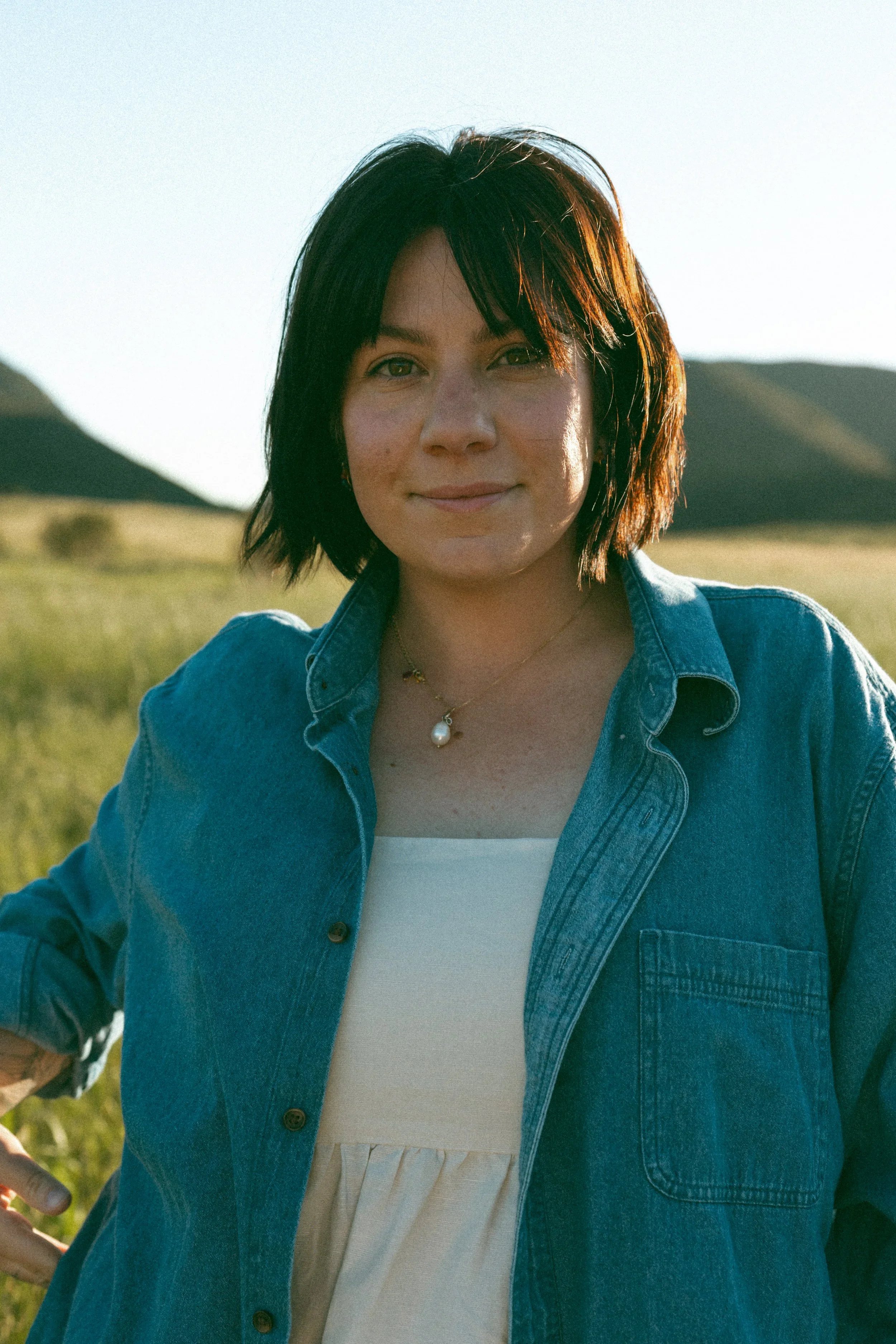 A woman with short dark hair standing outdoors in a grassy field with hills in the background, wearing a denim jacket over a white top and a pearl necklace.