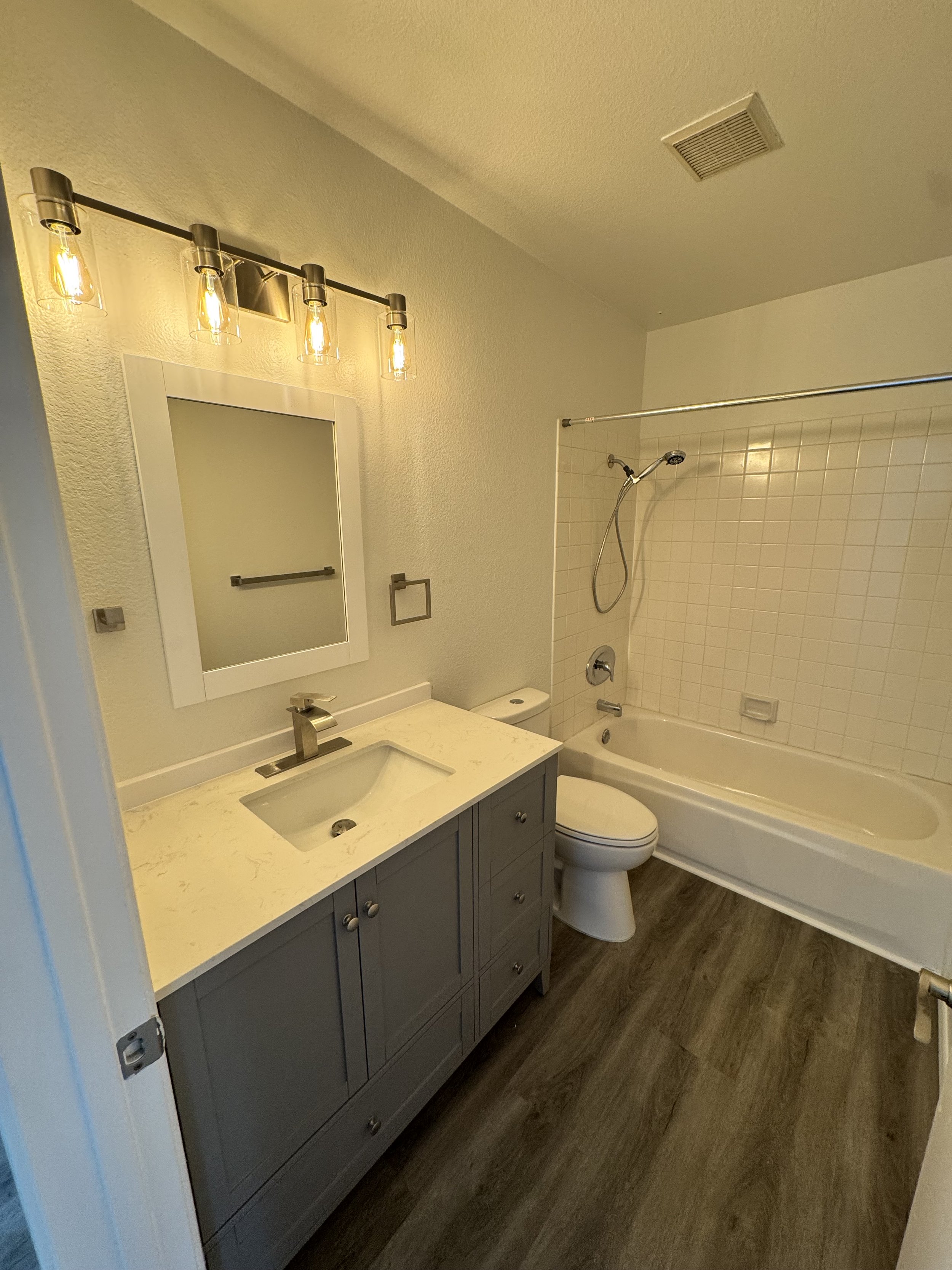 A bathroom with a vanity sink with beige marble countertop, a mirror, four exposed bulb light fixture, a toilet, and a bathtub with showerhead, all with wood-look flooring.