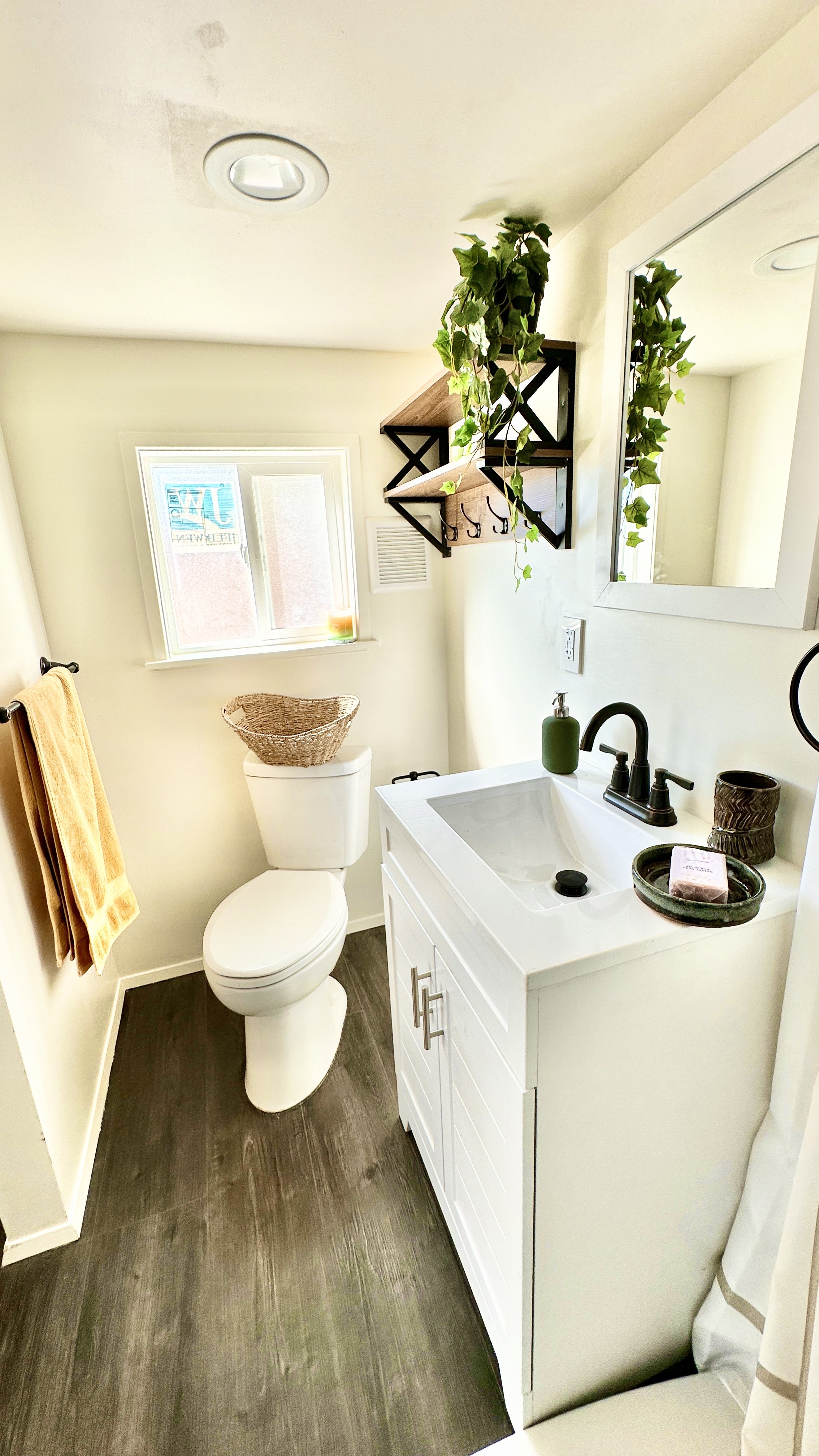 Small bathroom with white toilet, white vanity with sink, mirror, window, wall-mounted shelf with plants, yellow towels, dark wood floor.