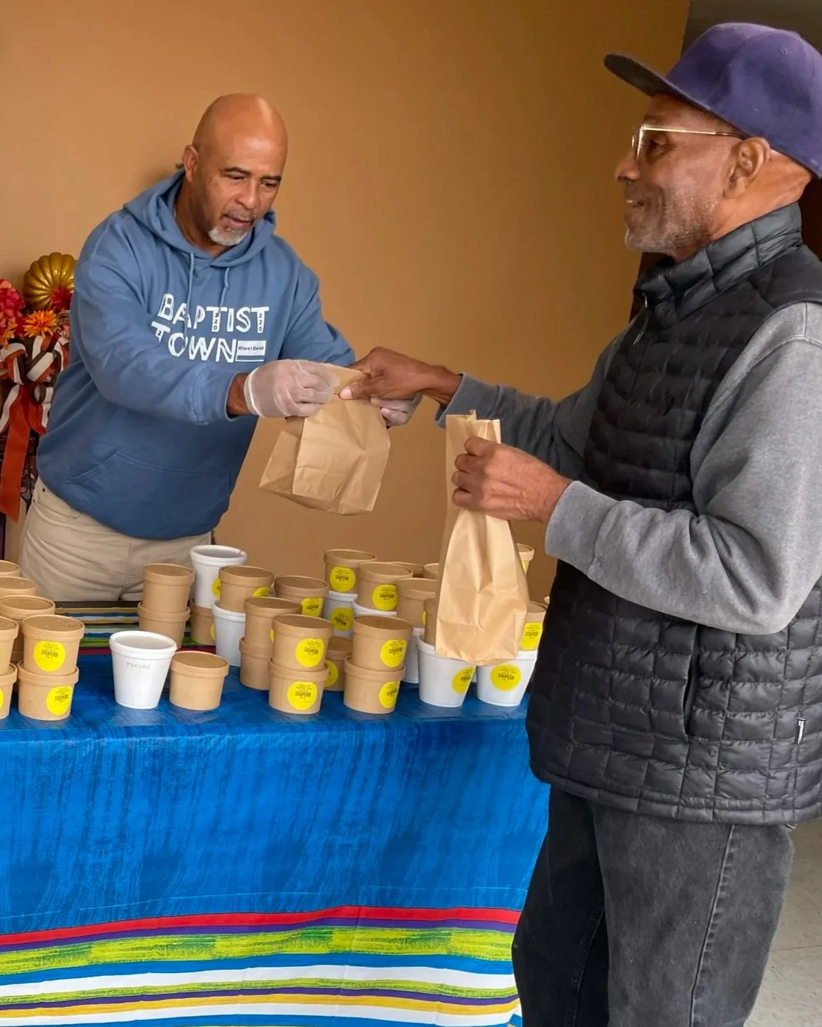 A man in a gray jacket and blue cap receives a brown paper bag from another man in a blue hoodie at a table with multiple paper and foam cups, some with yellow stickers, in what appears to be a food giveaway event.