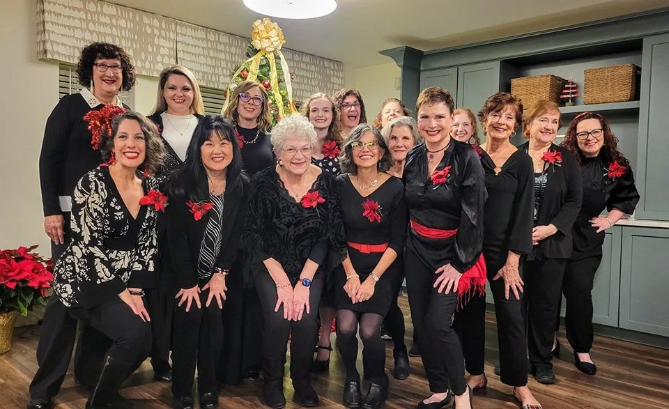 Group of singers dressed in black celebrating the winter holidays, with a wreath and poinsettias in the background.