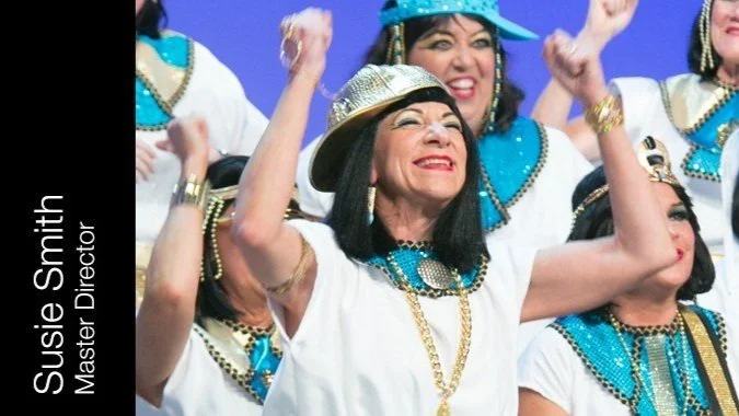 A group of singers dressed in white costumes with blue and gold accessories, celebrating on stage. The woman in front is wearing a silver hat, smiling and raising her arms.