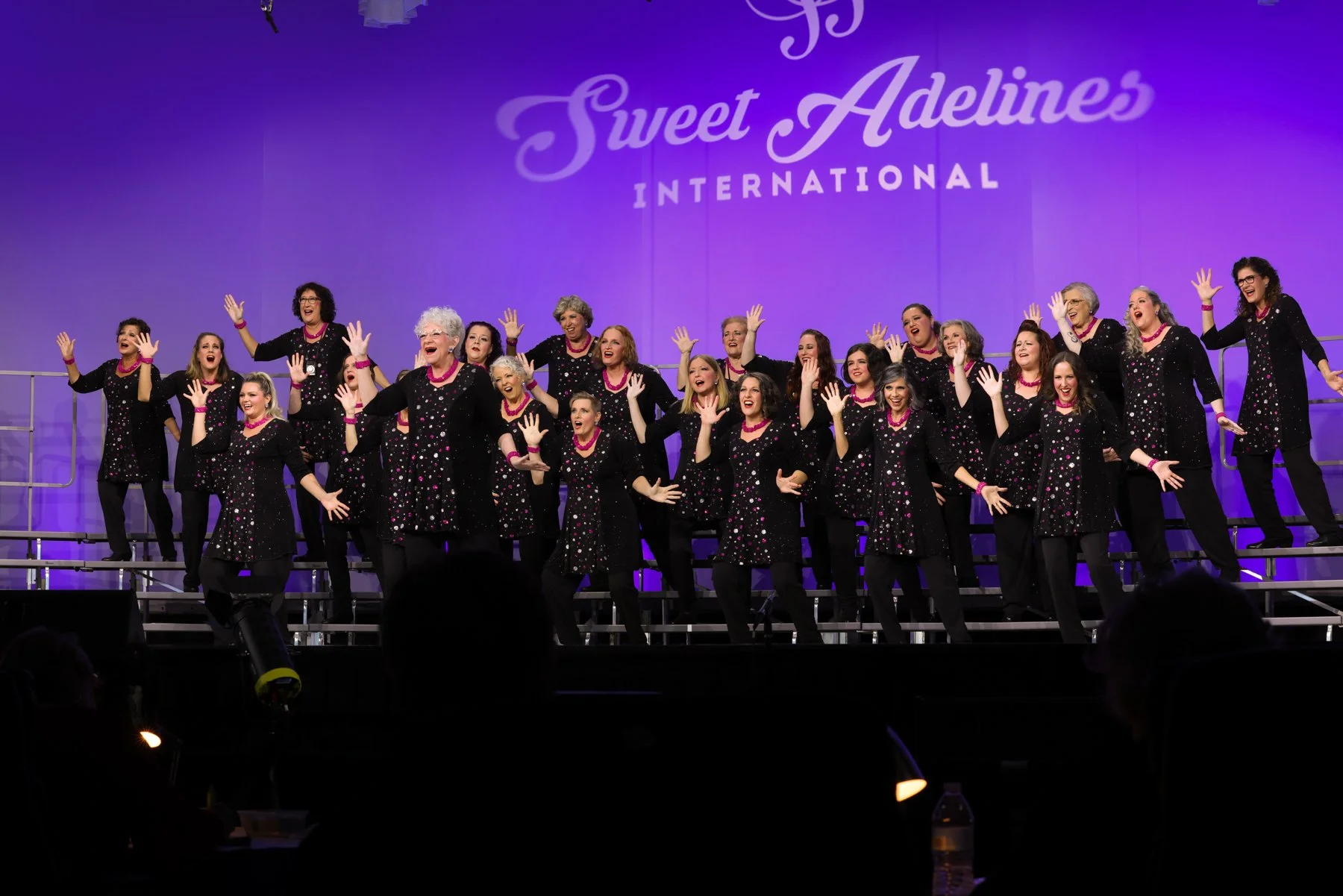 Group of  singers performing on stage at the Sweet Adelines International event, wearing matching black dresses with colorful polka dots, singing with smiles, in front of a purple backdrop.