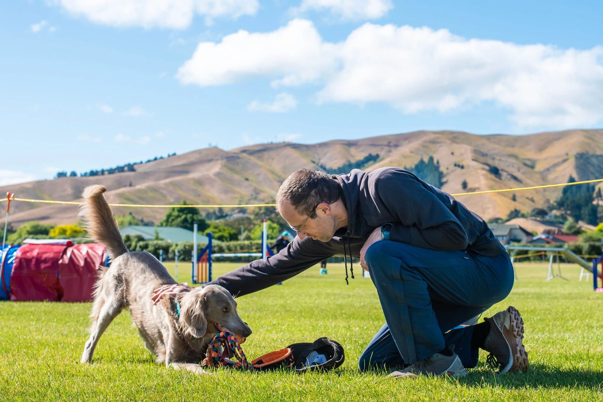 A man in a black hoodie and dark pants crouches on the grass, playing with a golden retriever puppy that has a colorful rope toy in its mouth, outdoors under a blue sky with clouds, with hills and a field in the background.