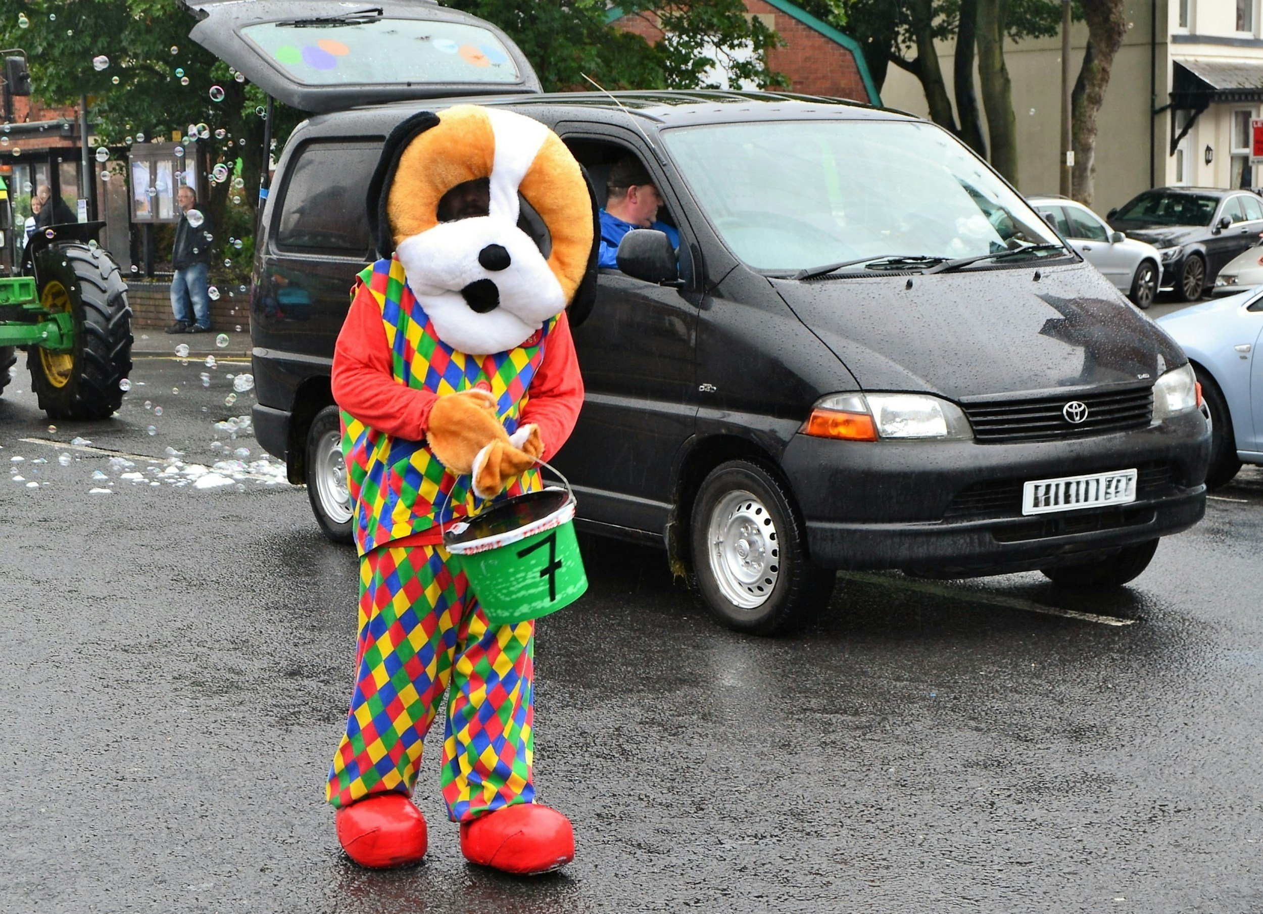 Person dressed in a colorful clown costume with a dog mascot head, holding a green bucket, standing on wet street with black minivan and parked cars in background.