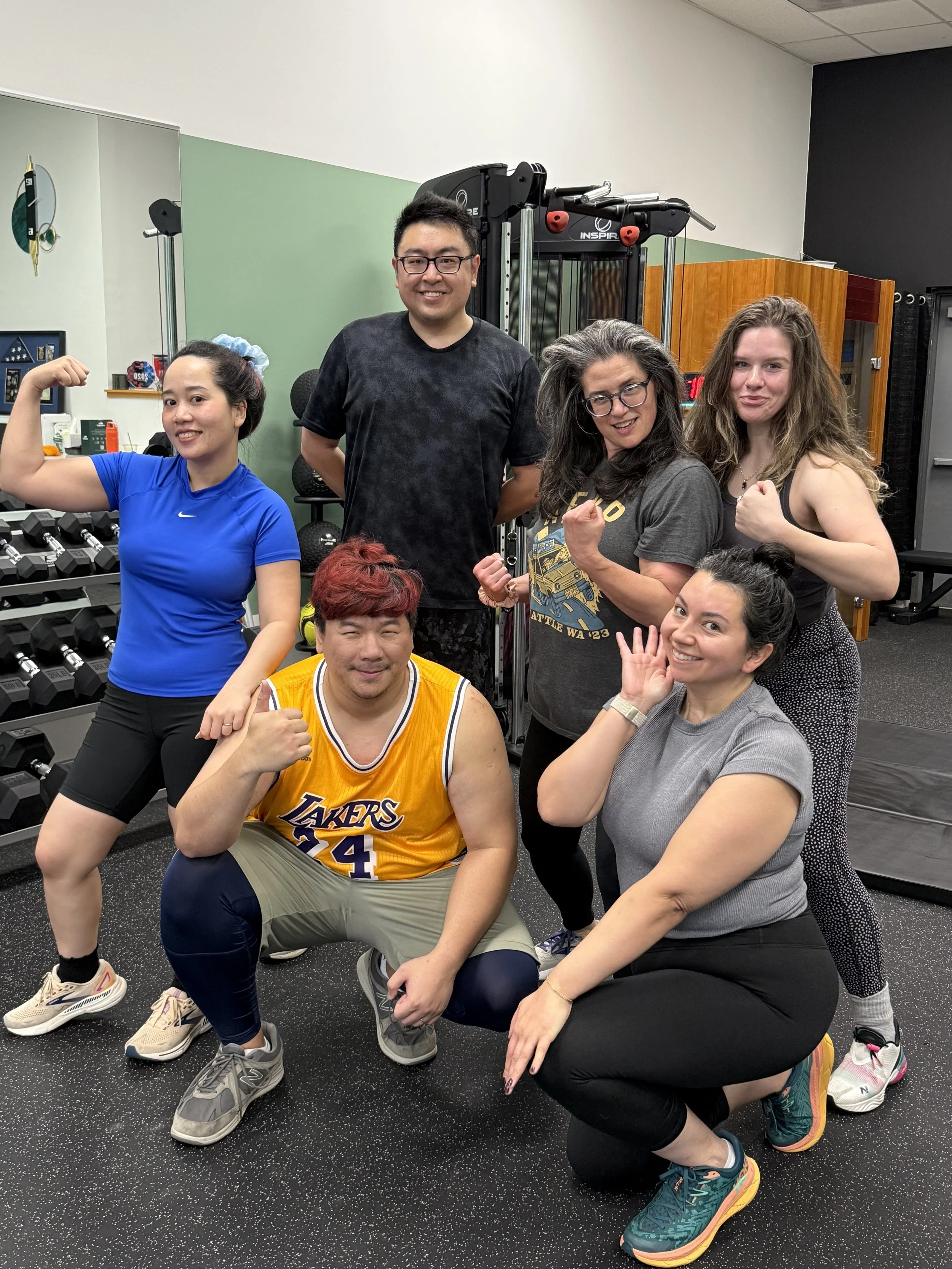 A diverse group of six people in workout clothes posing confidently in a gym, with some showing muscle and others making fist or peace signs, surrounded by gym equipment including dumbbells and exercise machines.
