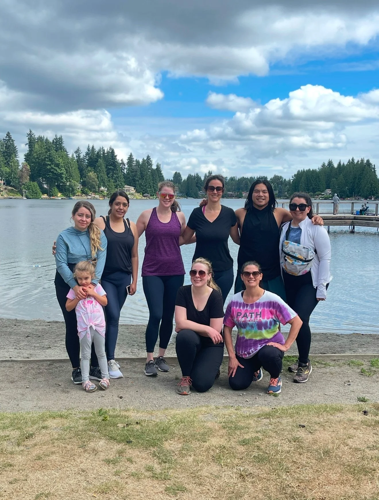 Group of eight women and one young girl posing by a lake with trees, clouds, and a cloudy sky in the background, some women wearing sunglasses and athletic clothing, smiling for the photo.