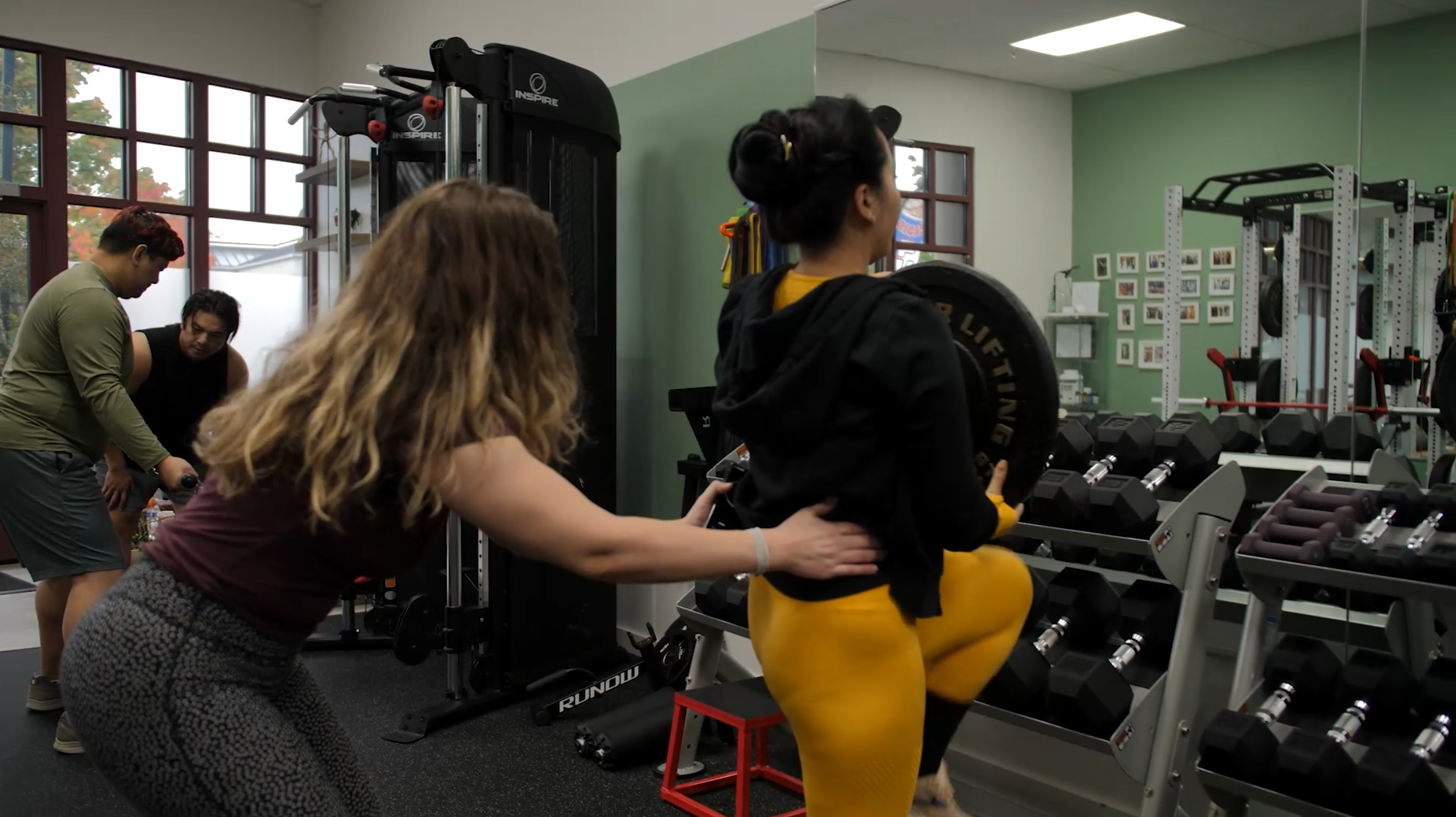 Woman in yellow leggings doing step-up exercise with weight on her shoulders in a gym, with trainer assisting her, and other people working out in the background.