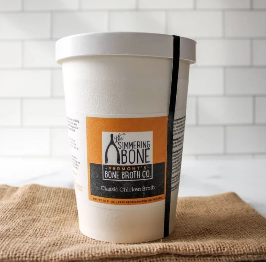 A container of bone broth, placed on a textured cloth against a white tiled background.