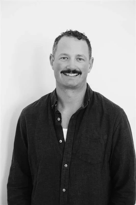 Black-and-white portrait of a smiling man with a mustache and short curly hair, wearing a dark button-up shirt over a light t-shirt, standing against a plain light-colored background.