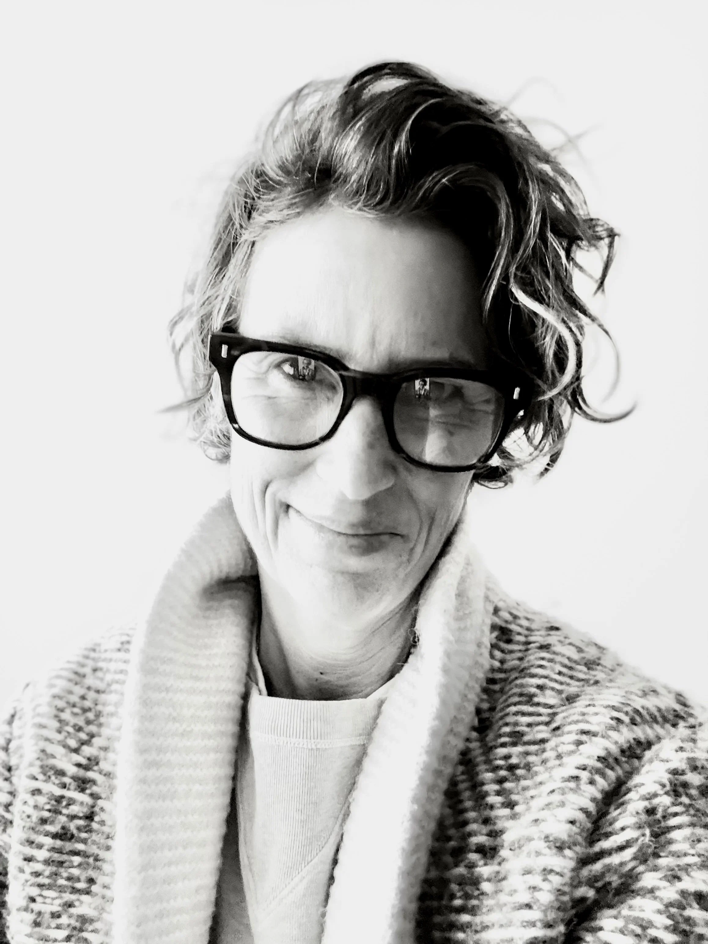 Black-and-white photo of a person with wavy hair, glasses, a smile, wearing a knit sweater and a t-shirt, against a plain background.