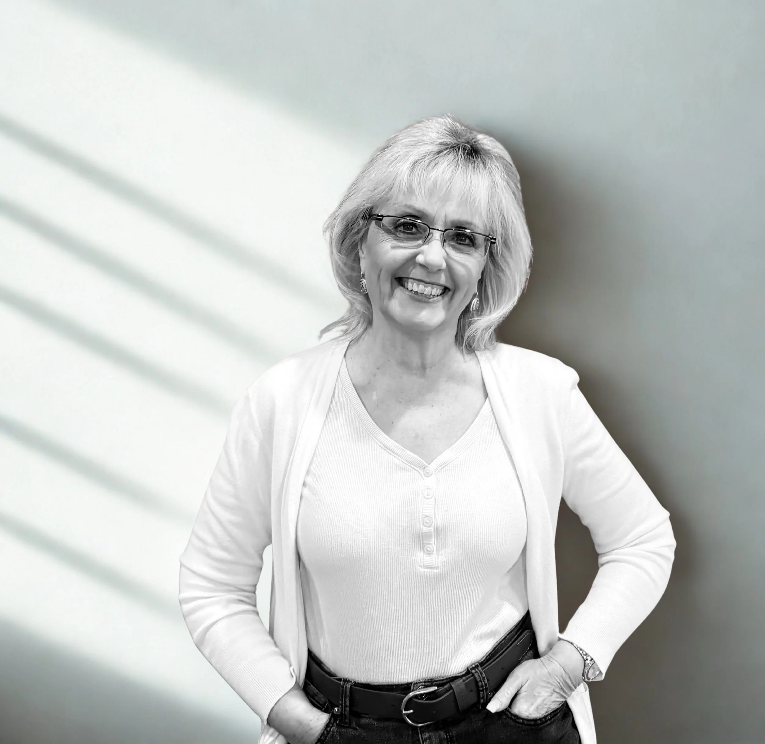 Smiling middle-aged woman with glasses, blonde hair, dressed in a white top and cardigan, standing against a plain wall with shadow lines.