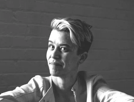 A woman taking a selfie against a brick wall, with short hair and a slight smile.