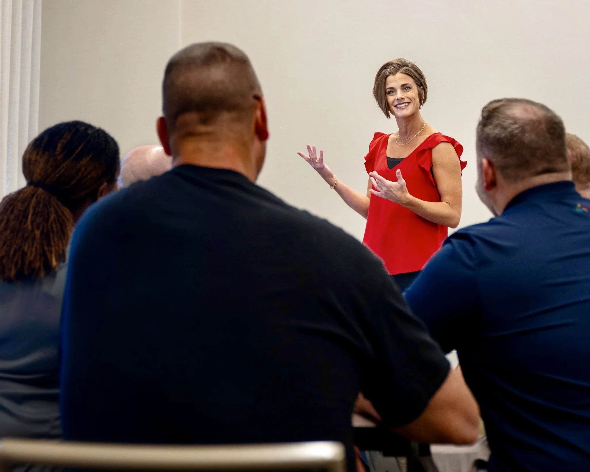 Executive coach Andrea Singsaas, wearing a bright red blouse, leads an interactive leadership development session, smiling and gesturing while engaging a small group of seated professionals.