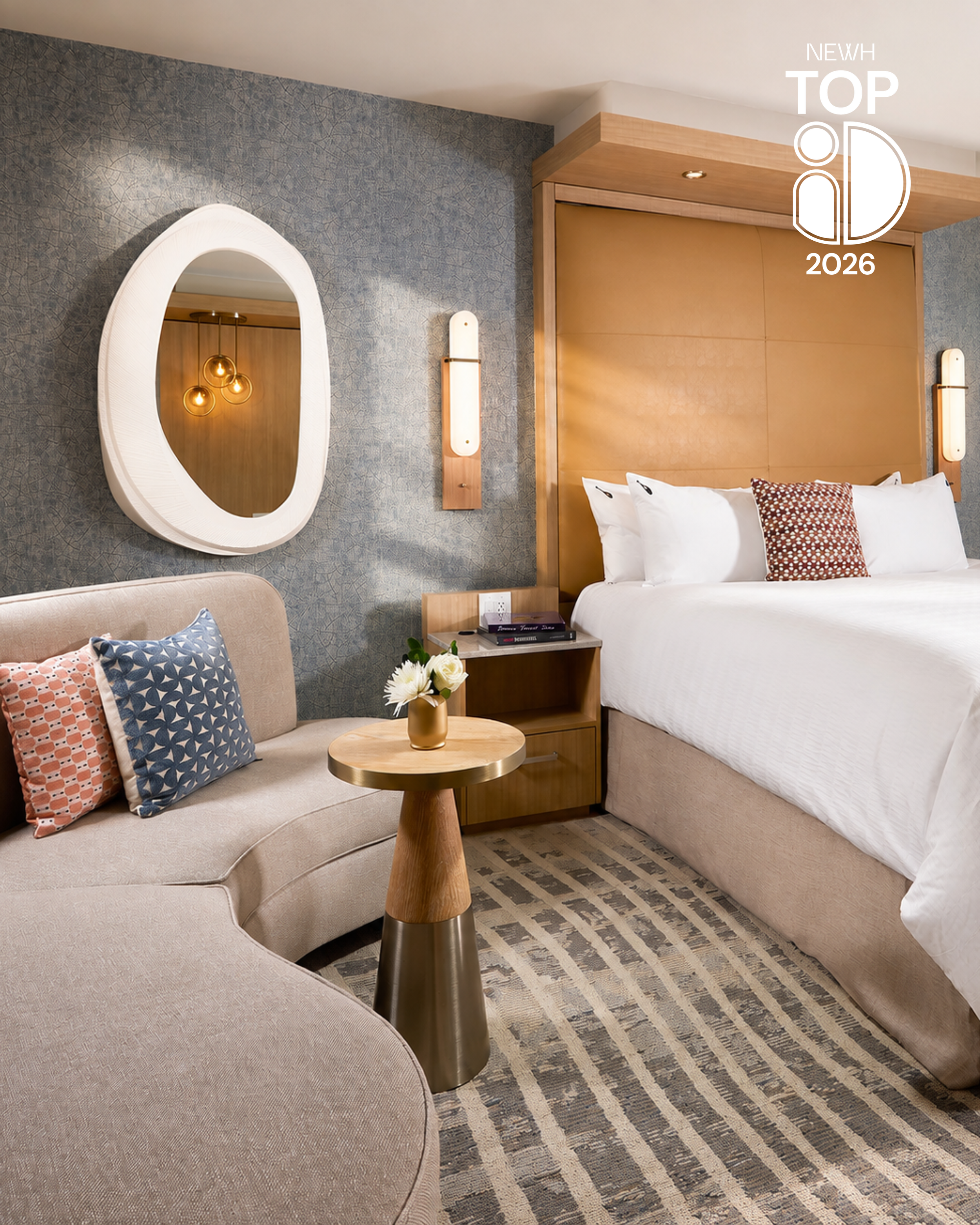 A hotel room with a beige sofa with three decorative pillows, a small round wooden table with a flower vase, a bed with white linens and a patterned pillow, wooden headboards, wall-mounted lamps, a mirror with a white frame, textured gray wallpaper, and a patterned carpet.
