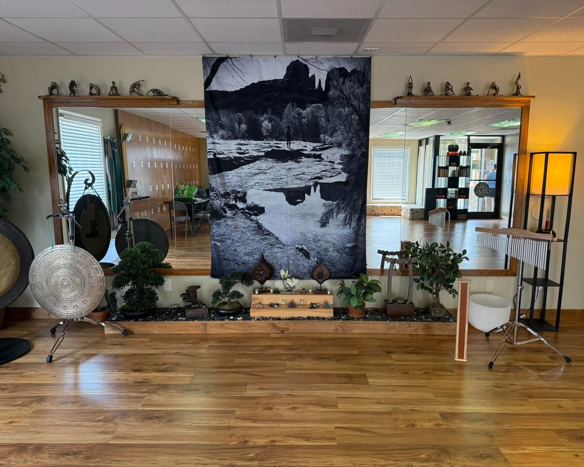 Indoor room with wooden floor, plants, Tibetan singing bowls, gong, marimba, and a large black-and-white landscape tapestry of a river and mountains hanging between two mirror walls reflecting furniture and windows.