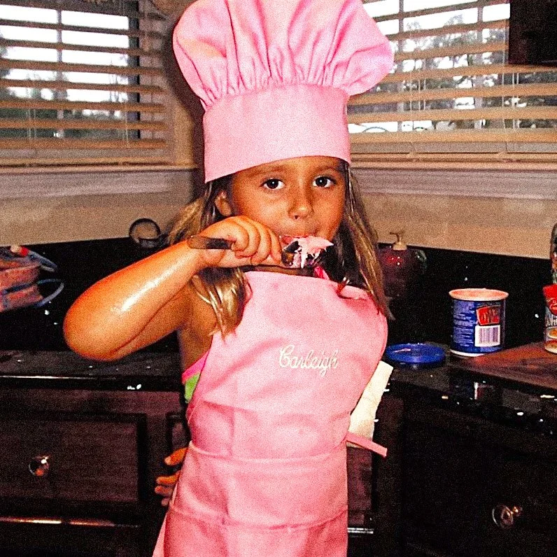 A young girl wearing a pink chef hat and pink apron, eating a piece of cake with a fork in a kitchen.