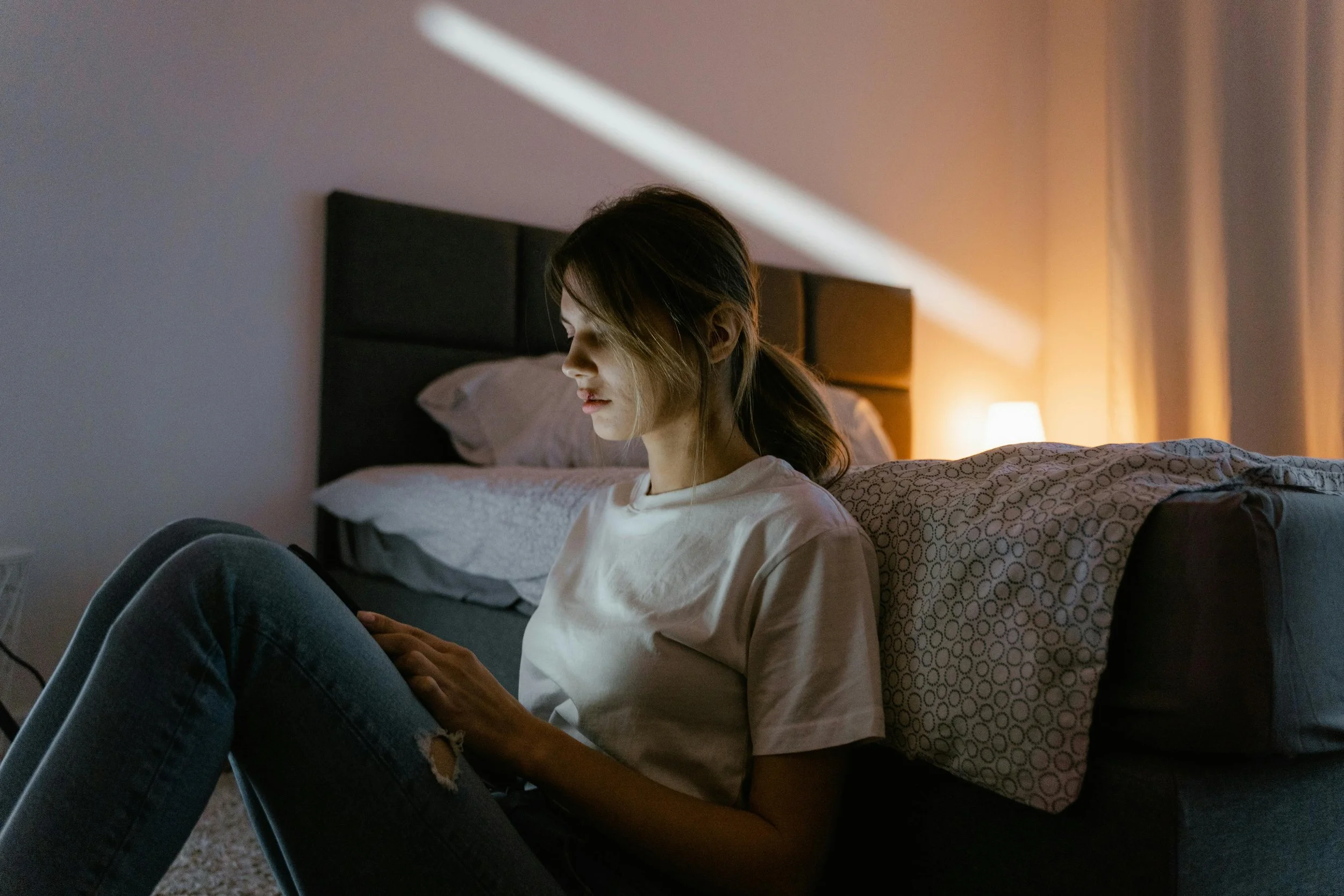 A woman sitting on the floor leaning against a bed in low light
