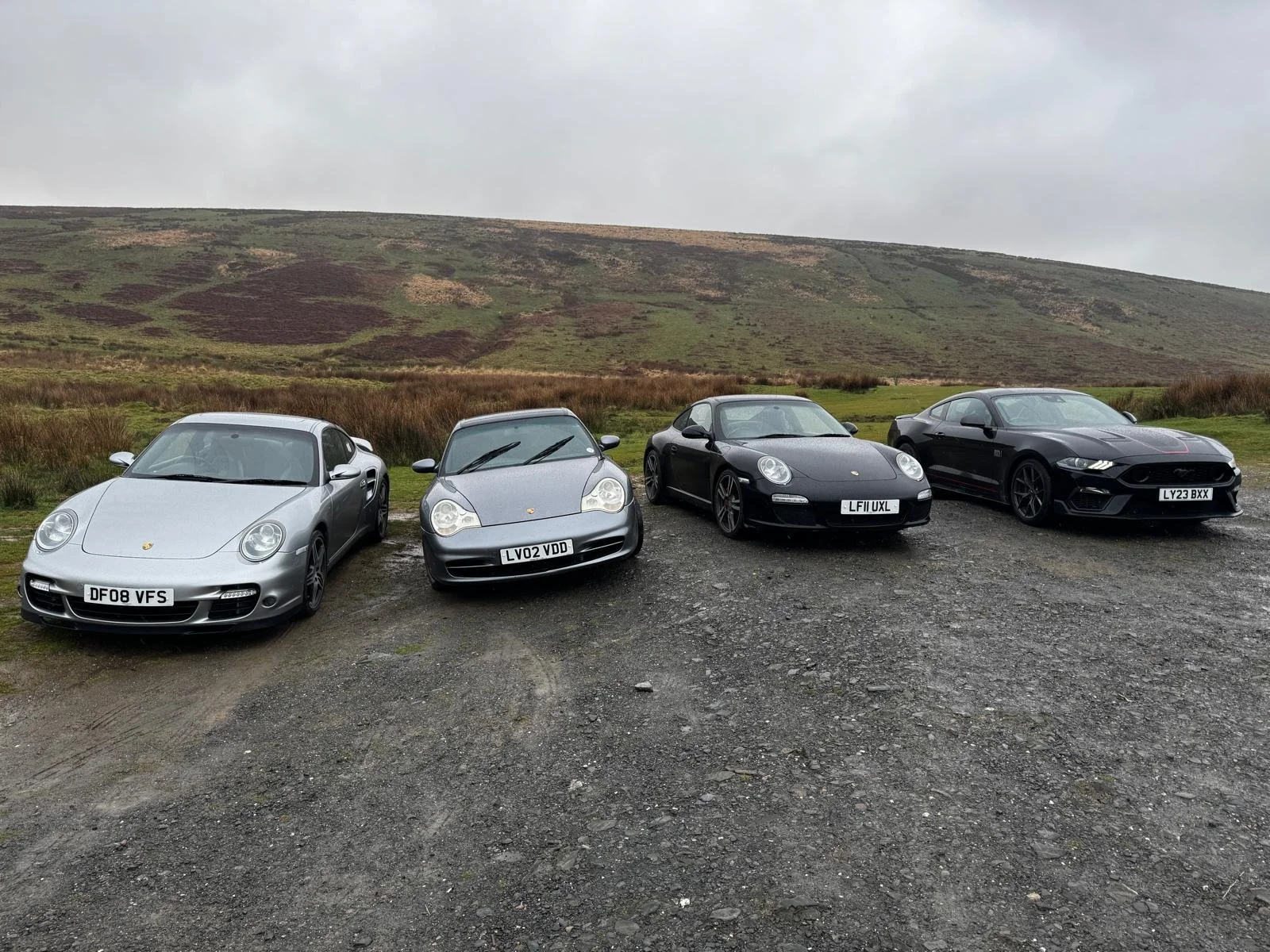 Four Porsche sports cars parked on a gravel surface in front of a grassy hillside and cloudy sky. The cars are silver, dark gray, black, and dark gray with red accents, from left to right. On a UK Road Trip