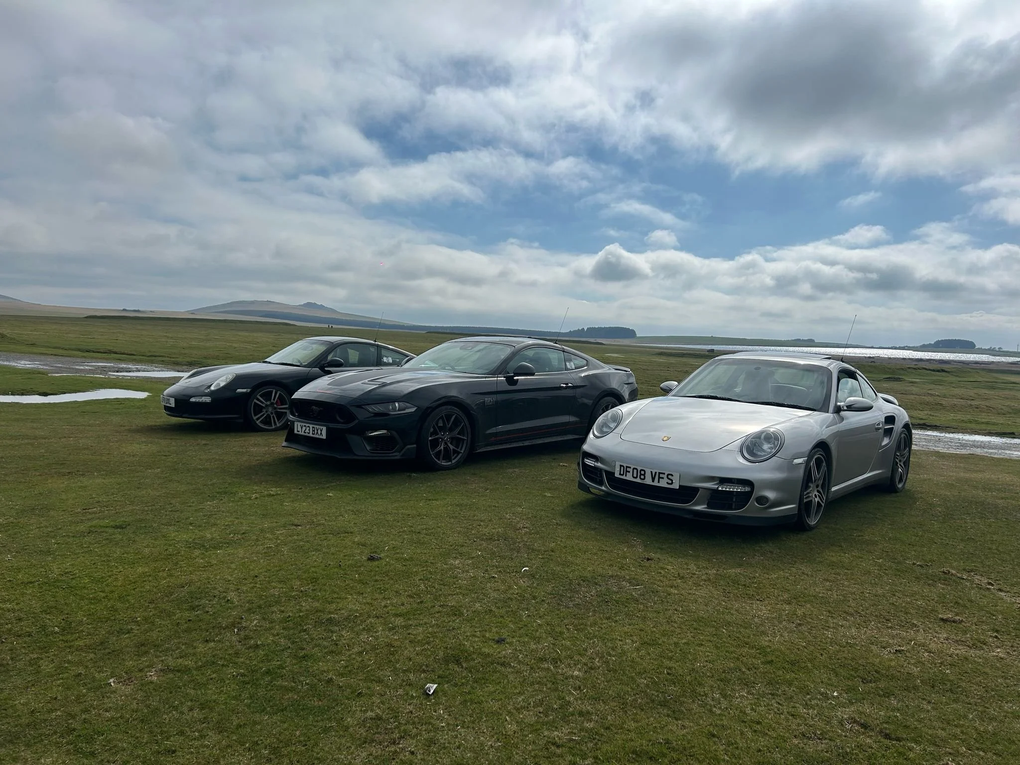 Three sports cars parked on a grassy field under a partly cloudy sky with a lake and rolling hills in the background. From left to right, a black Porsche, a black Mustang Mach 1, and a silver Porsche 911.