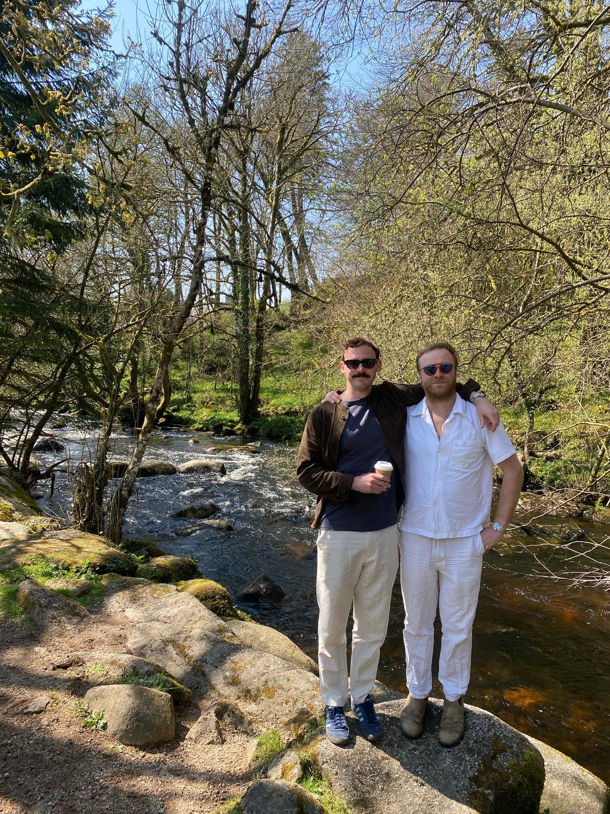 Two men standing on rocks by a river in a wooded area, with one holding a coffee cup, both wearing sunglasses and casual clothing, under a clear sky. Enjoying a break on a UK Road Trip in beautiful surroundings