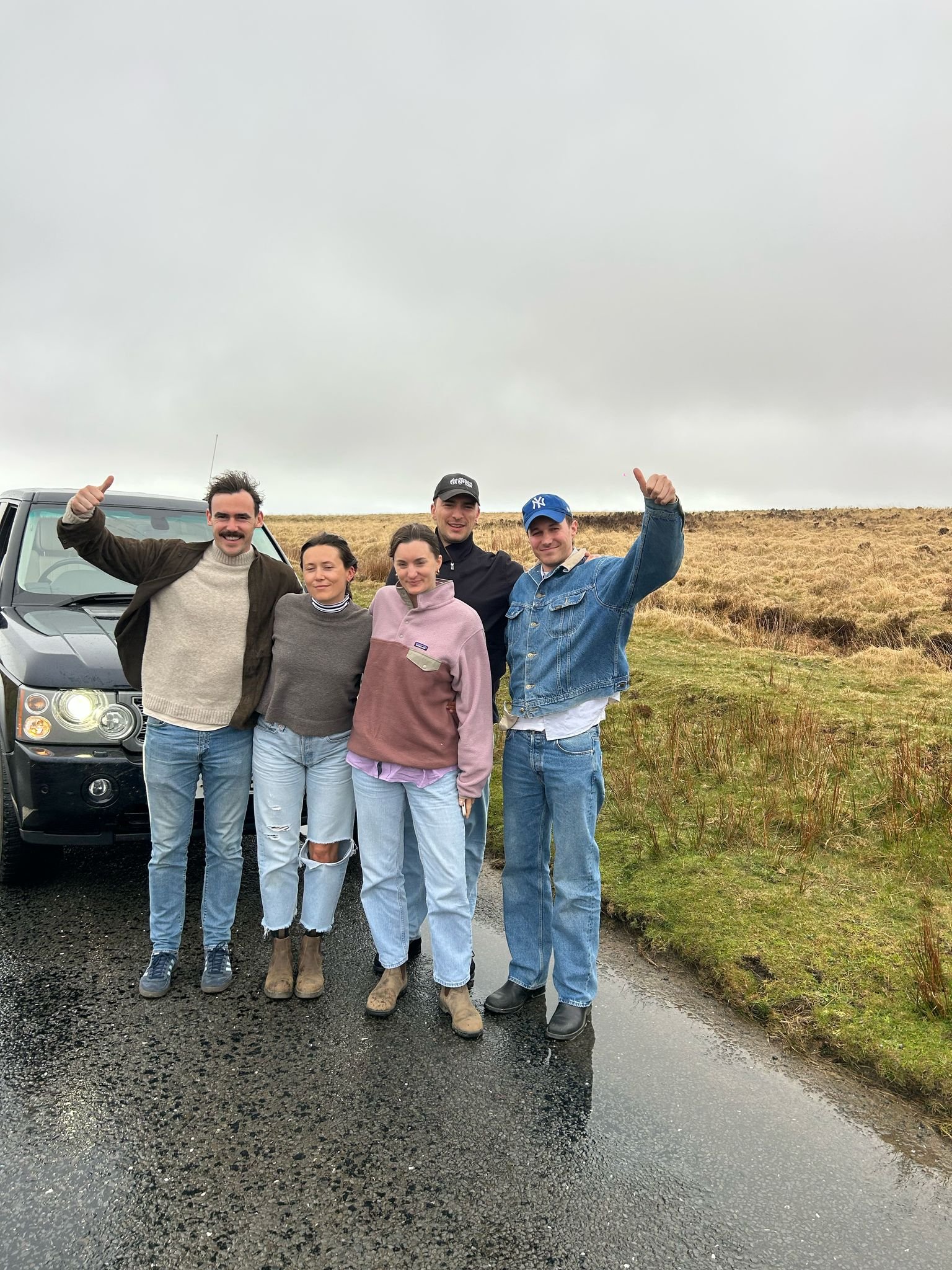 Group of five friends standing outside on a wet road with a vehicle behind them, smiling and posing with thumbs up, in a grassy, hilly area under an overcast sky. Enjoying a UK Road Trip in Exmoor