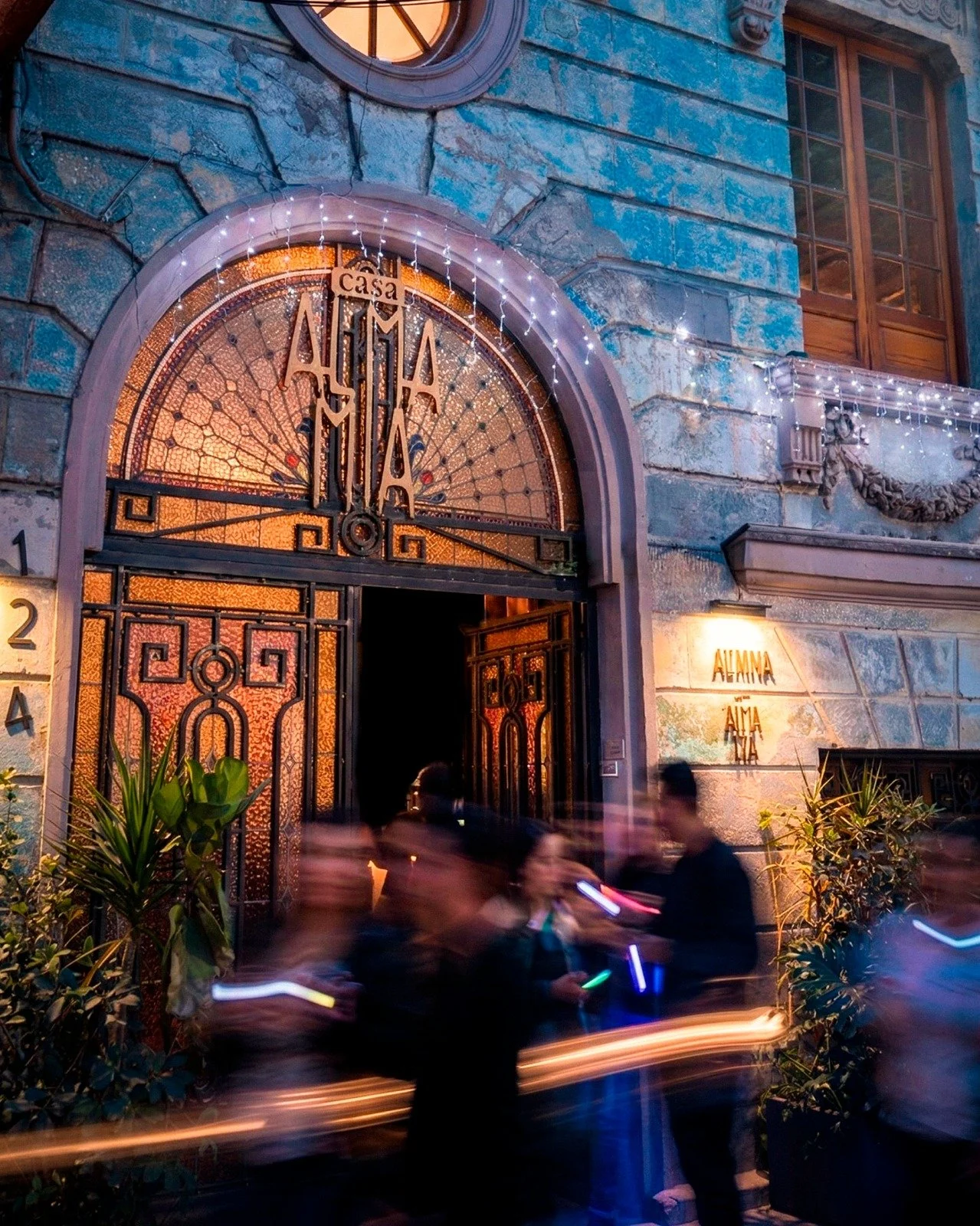 Puerta de hierro forjado con un arco de piedra y una ventana circular en la pared de una casa antigua, con enredaderas y cables eléctricos.
