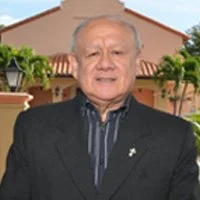 An elderly man in a black suit standing outdoors with a building and greenery in the background.