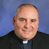 Portrait of a smiling clergyman in a black suit and clerical collar against a blue background.