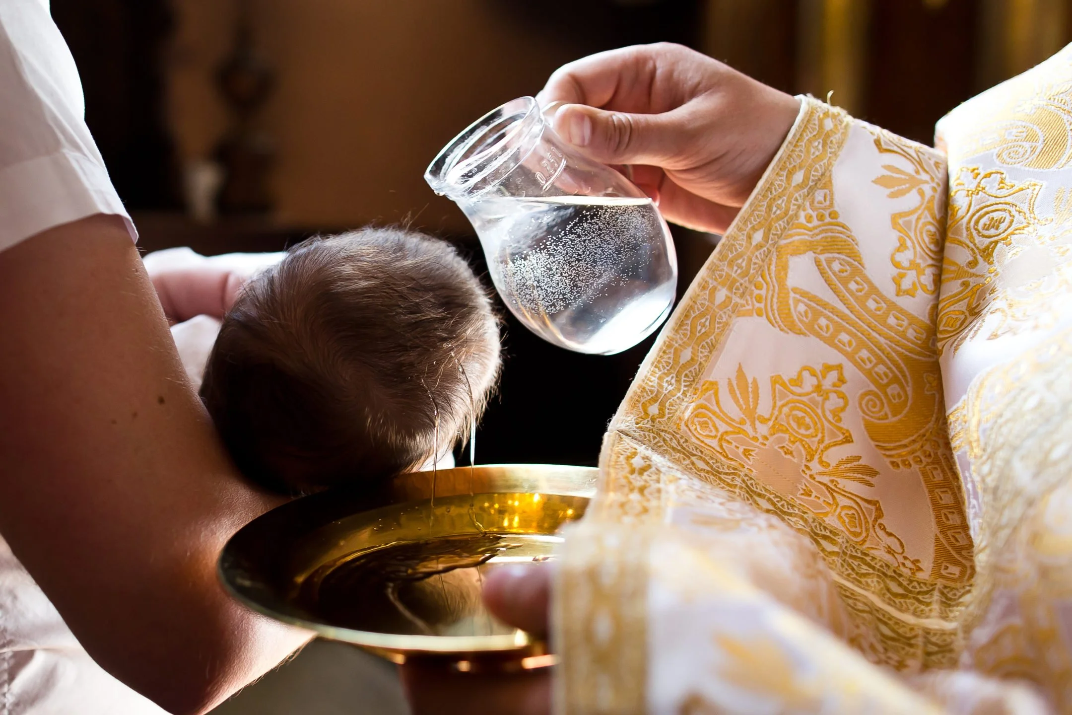 A priest in ornate gold and white robes blesses a baby with holy water from a glass vessel held in his right hand, while the baby's mother holds the baby. The baby's head is bowed as water is poured over it.