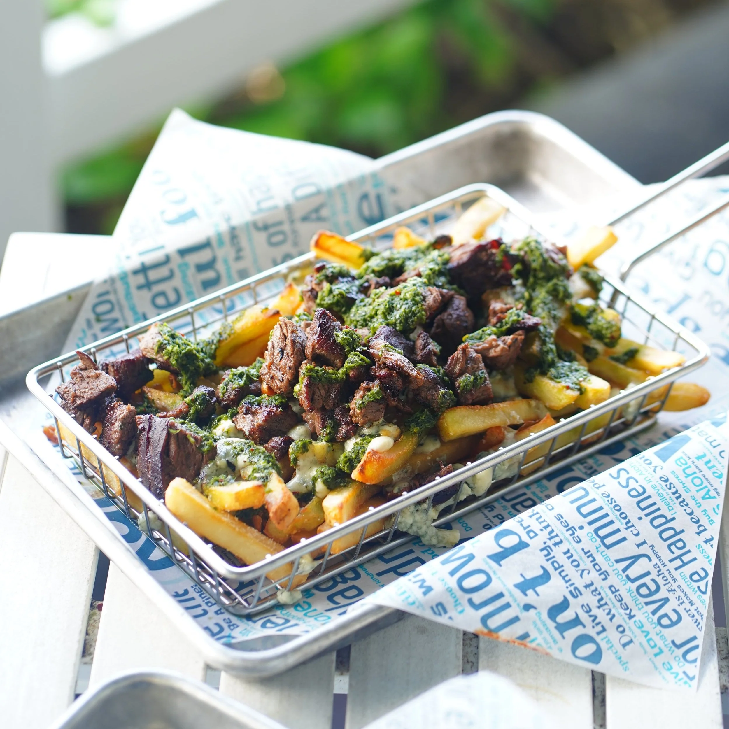 French fries topped with sliced beef and green sauce, served in a metal basket lined with colorful branded paper.