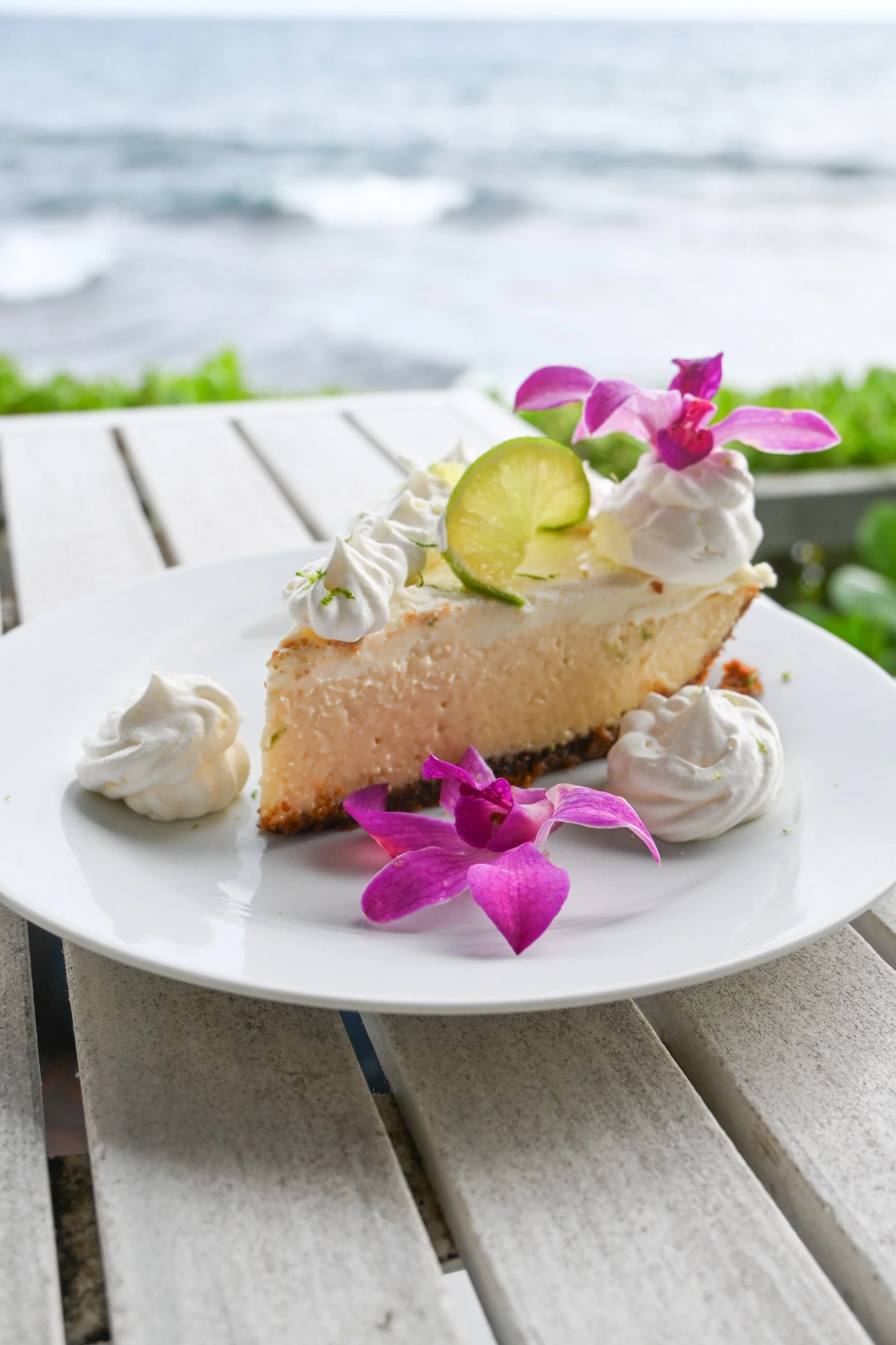 Slice of cheesecake garnished with lime slice, whipped cream, and purple orchids on a white plate, with ocean view in the background.