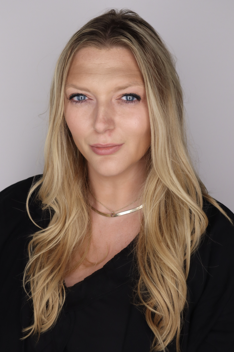 A portrait of a woman with long blonde hair, blue eyes, and a subtle smile, wearing a black top and layered gold necklaces, against a plain light background.