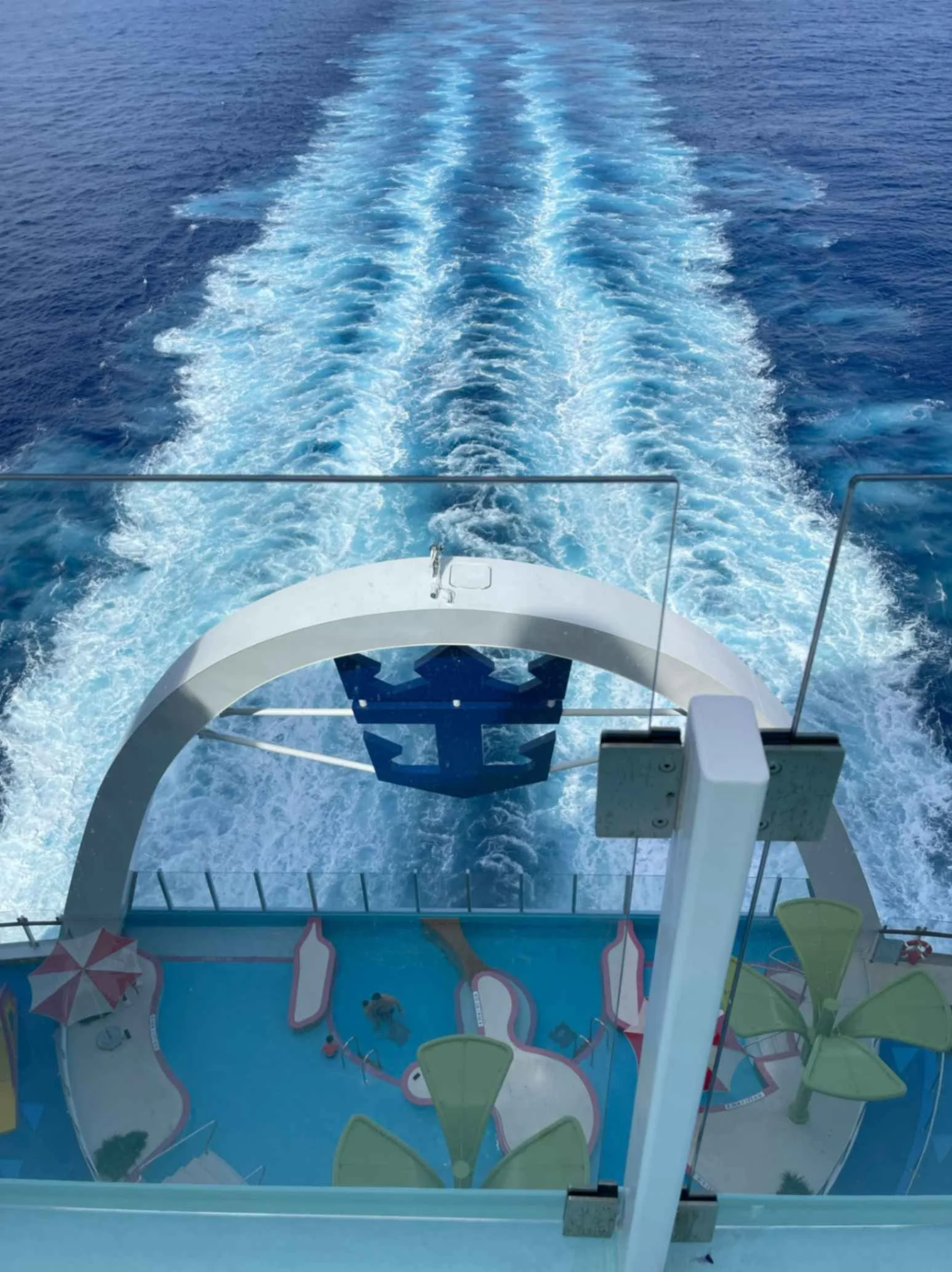View from the aft of Royal Caribbean’s Icon of the Seas showing the ship’s wake cutting through deep blue Caribbean waters. The Royal Caribbean Crown & Anchor emblem is visible above a splash zone and water play area, symbolizing adventure and luxury