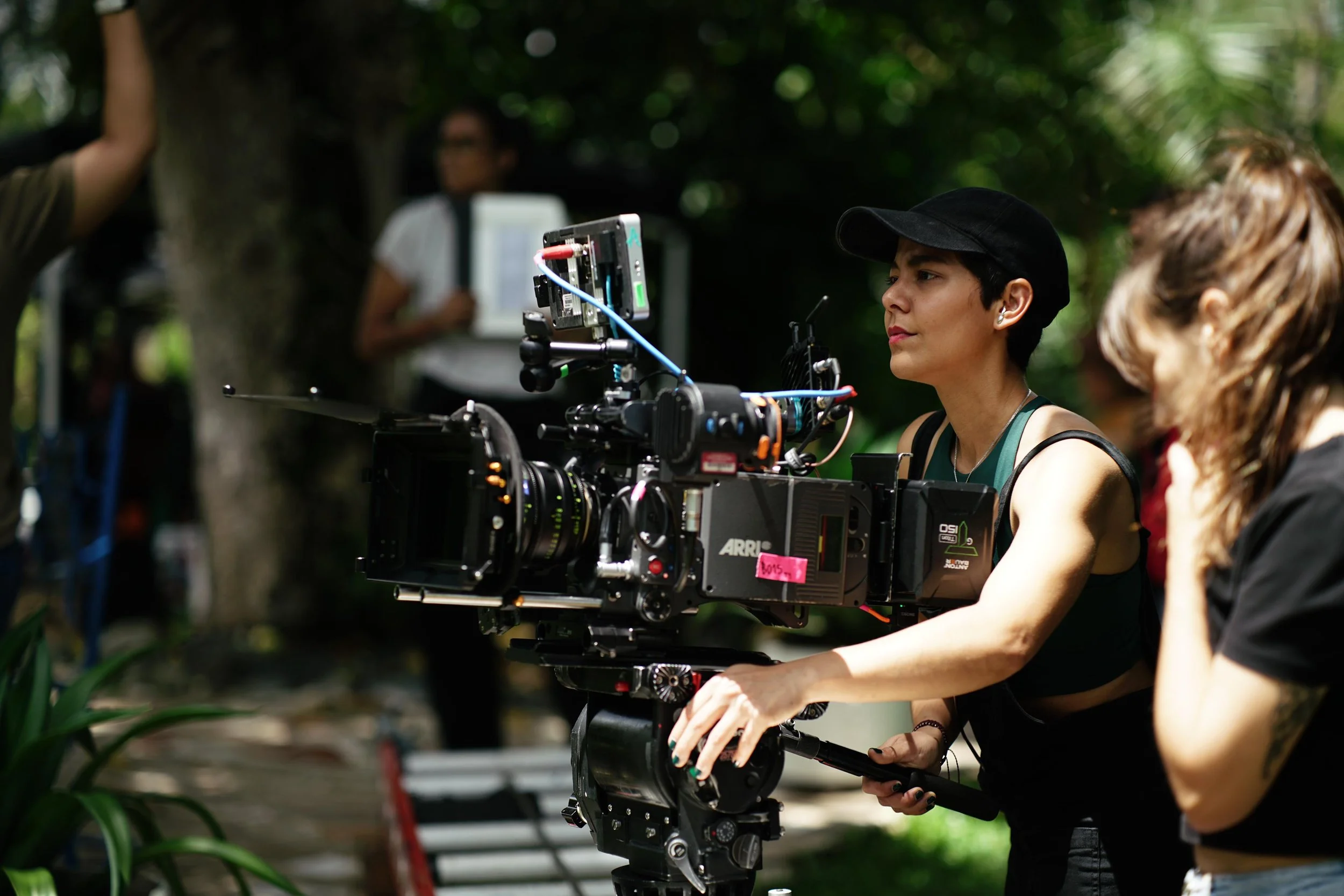 Miriam Ouchi operates a professional ARRI studio camera outdoors during a film shoot, with other crew members visible in the background.