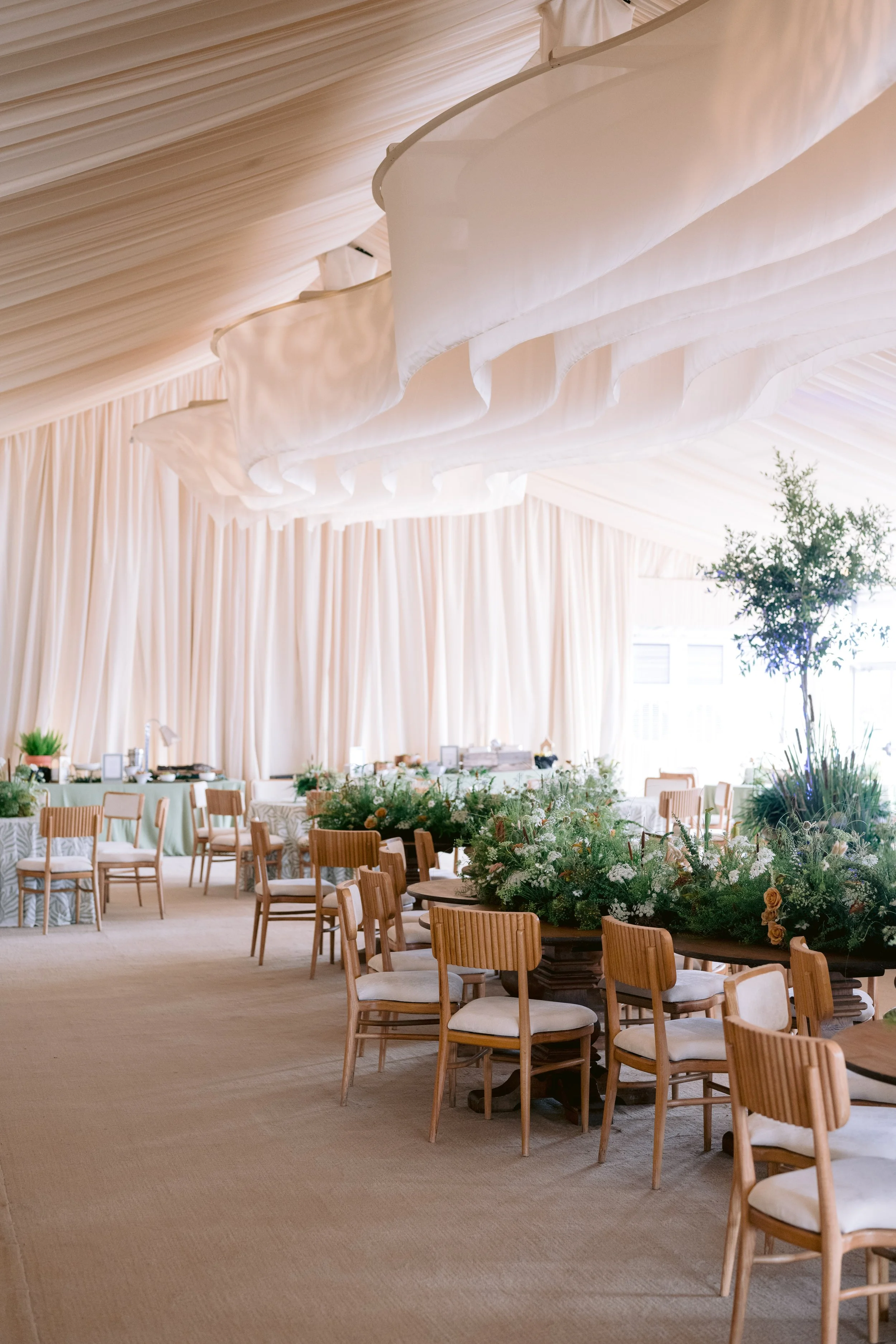 Elegant event space with round tables covered in floral centerpieces, surrounded by wooden chairs with white cushions, under a draped ceiling with soft fabric decoration and natural light filtering through windows.