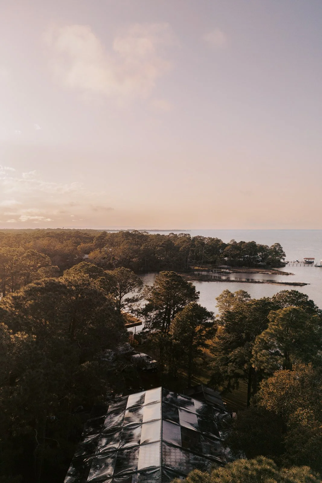 Aerial view showing a body of water surrounded by trees, with a dock and boats in the background, and rooftops of buildings in the foreground during sunset.