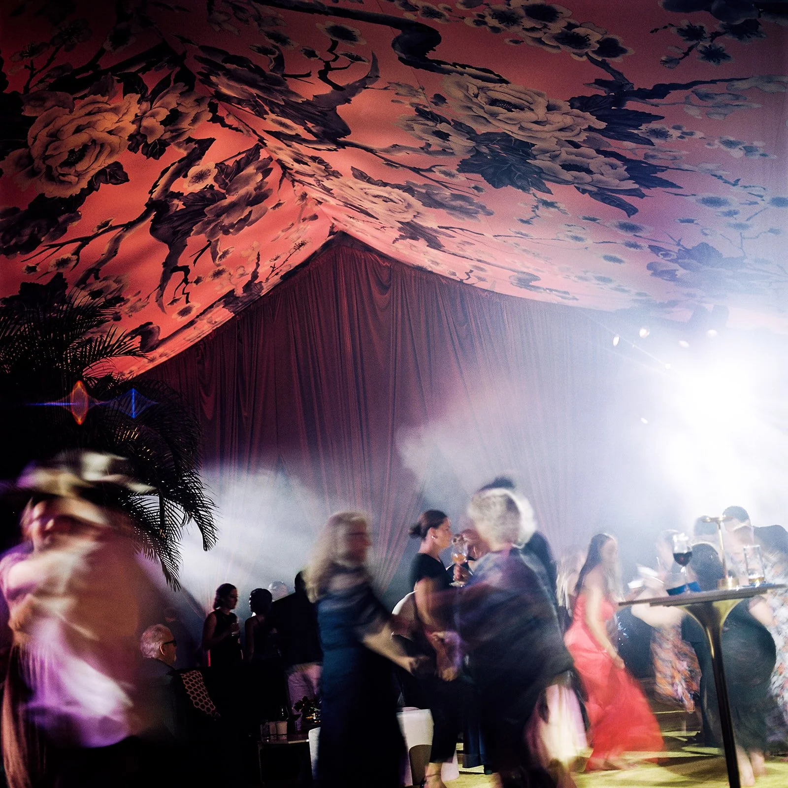 People dancing and socializing under a colorful, floral ceiling with bright lighting at a lively event or party.