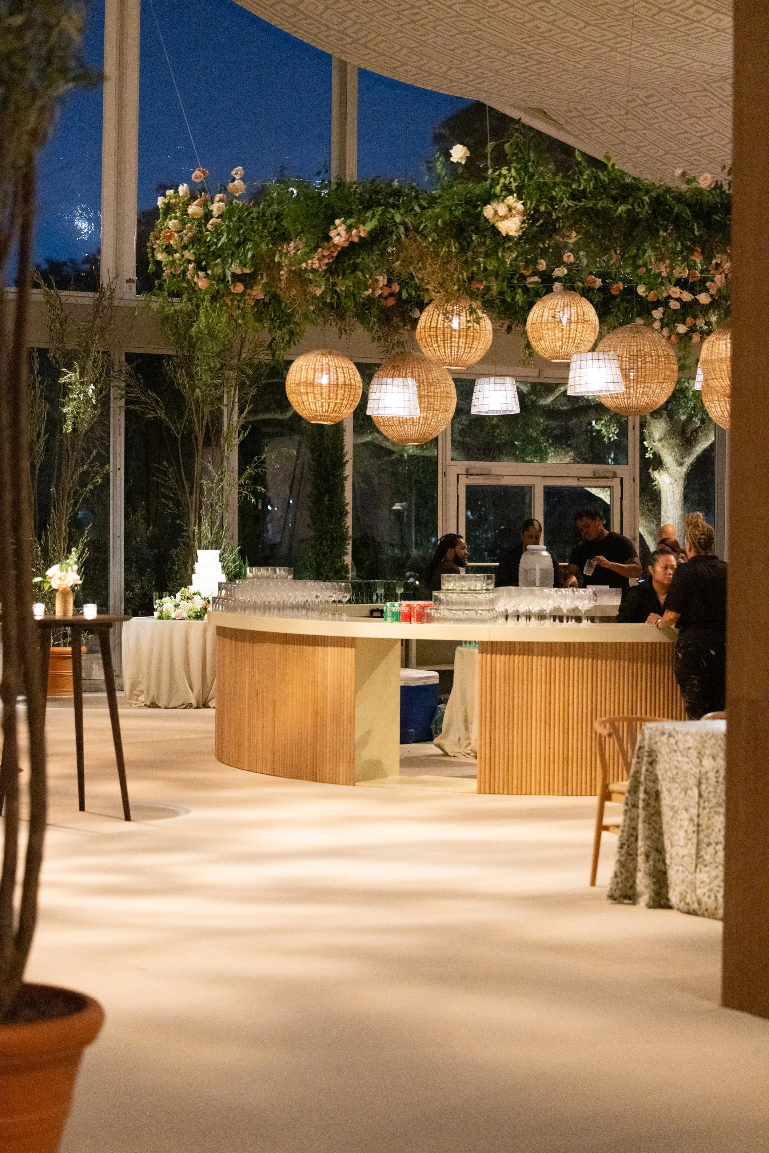 Elegant indoor reception area with hanging wicker and fabric light fixtures, floral arrangements, a curved wooden bar, and people preparing drinks at night.