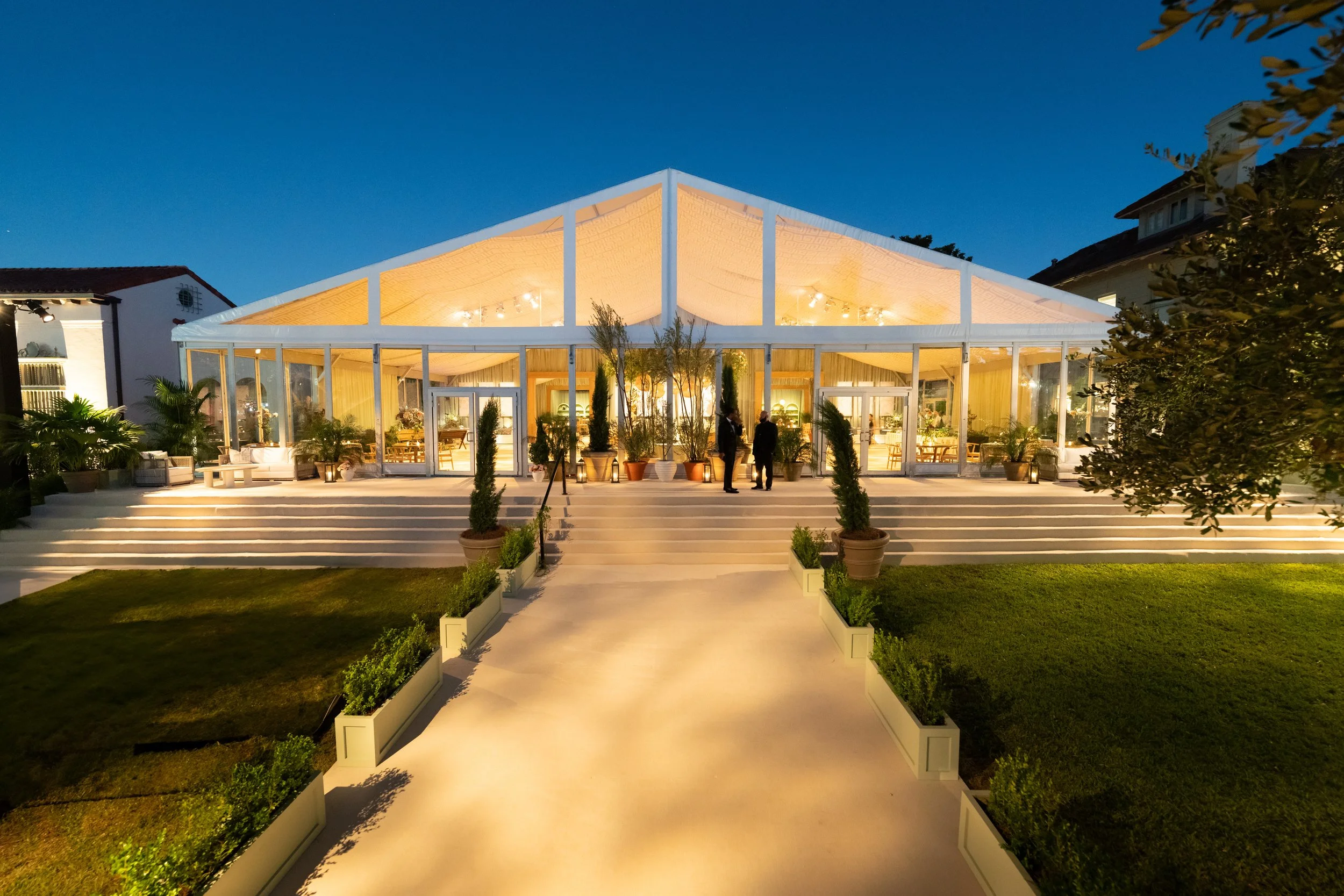 Elegant glass venue with warm lighting, surrounded by potted plants and a garden, set for an evening event.