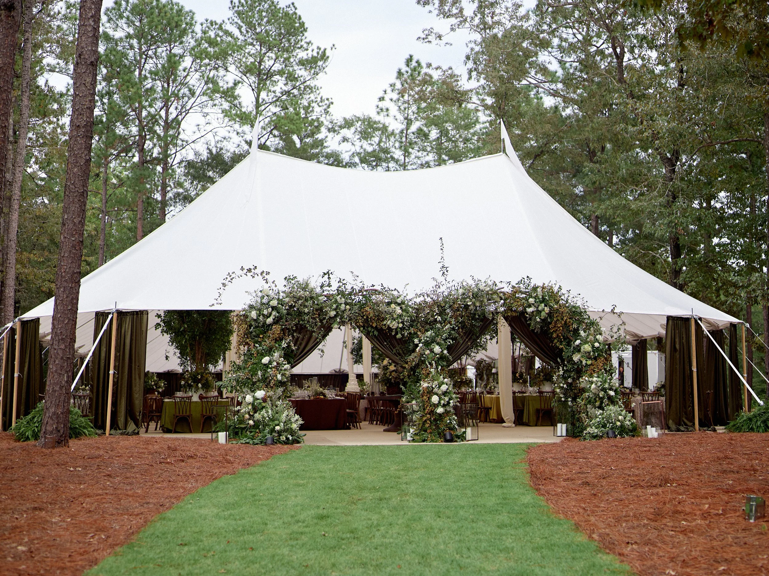 A large outdoor wedding tent decorated with white flowers and greenery, set up in a wooded area with a grassy pathway leading to it.
