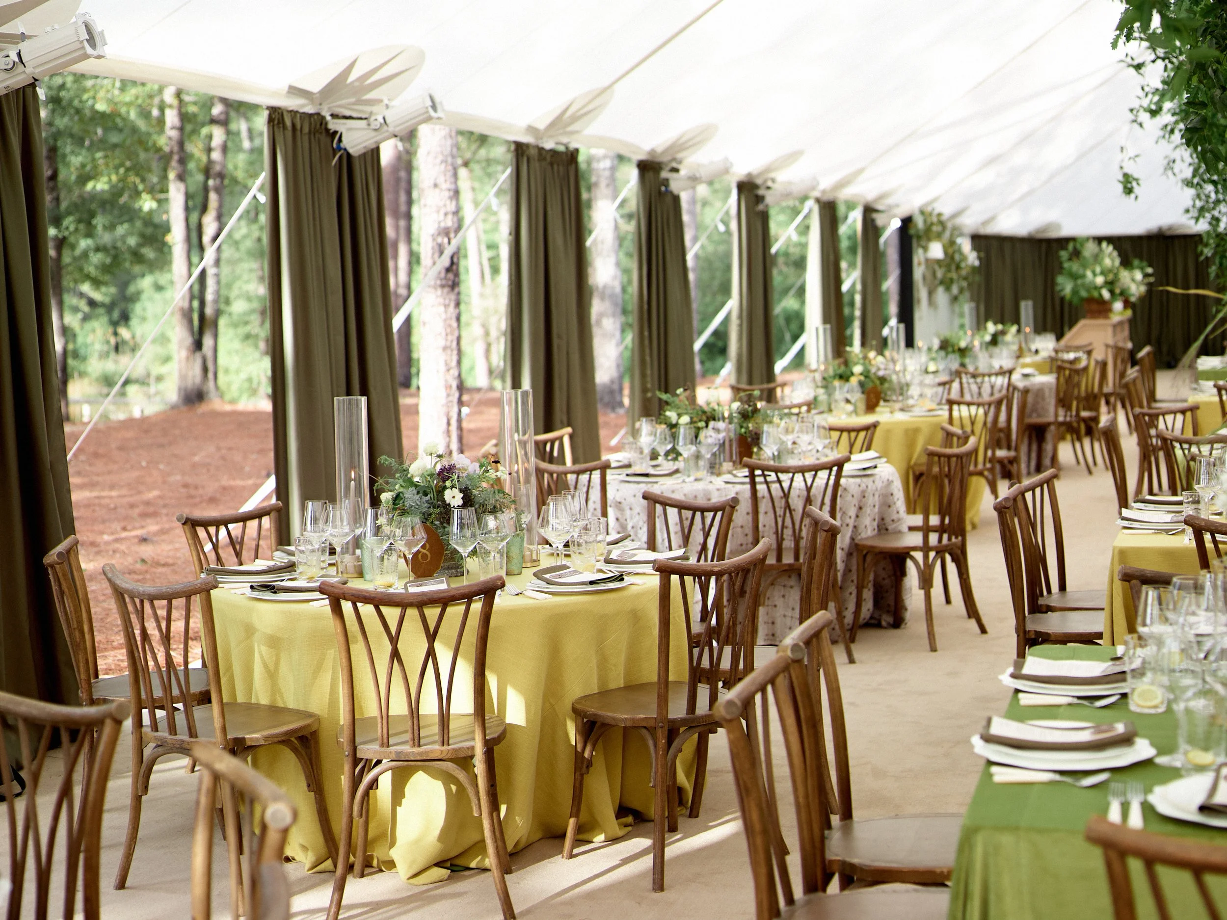 Reception tent with round tables covered in yellow, green, and patterned tablecloths, decorated with flower centerpieces, glassware, and place settings, surrounded by wooden chairs with trees visible outside.