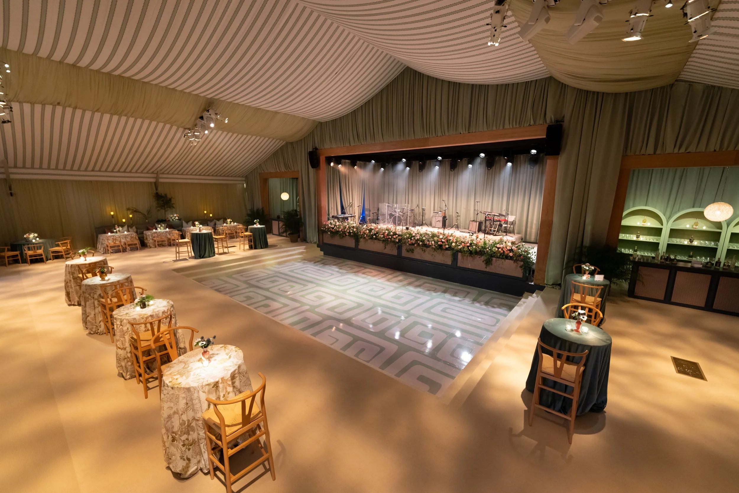 Elegant event space with a stage decorated with flowers, small round tables with floral centerpieces, and a dance floor, all set preparation for a formal gathering or celebration.