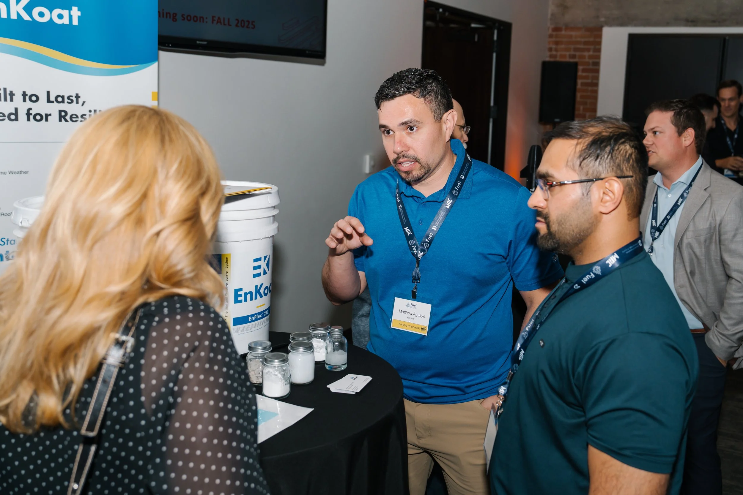 A man in a blue shirt is talking to a woman with red hair at a trade show or conference, with other attendees in the background. There are jars with powder or white substances on the table and a banner with the EnKoat logo.