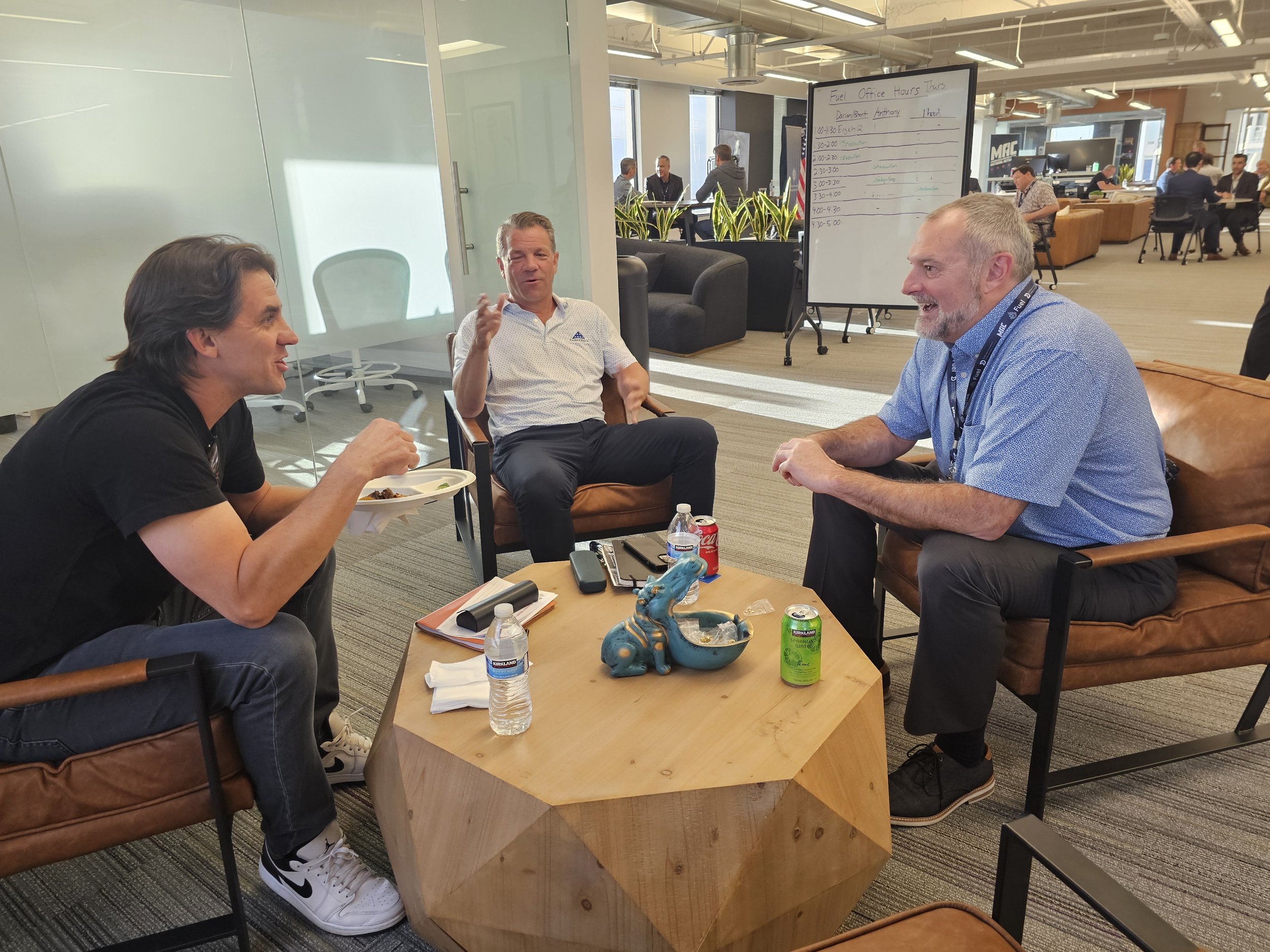 Three men sitting and conversing in an office lounge, two are seated on brown cushioned chairs while one is seated on a leather sofa. There is a small wooden table with drinks, snacks, and remote controls. In the background, there are other people wo