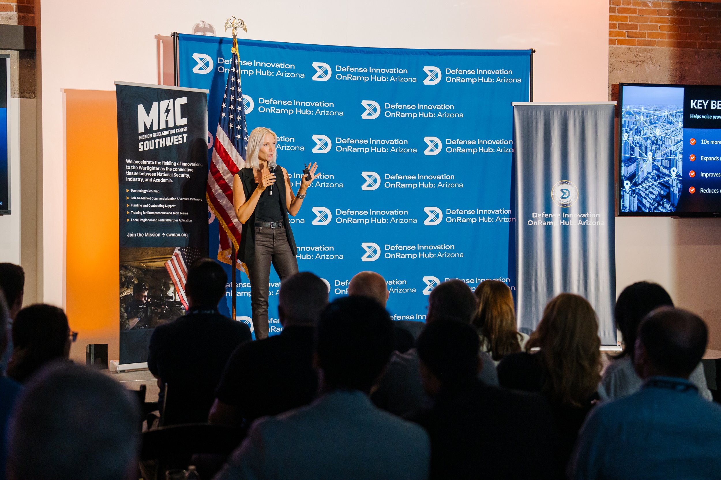 A woman giving a presentation on stage at the Defense Innovation OnRamp Hub in Arizona, with a large blue backdrop and a screen displaying information, an American flag, and an audience in attendance.