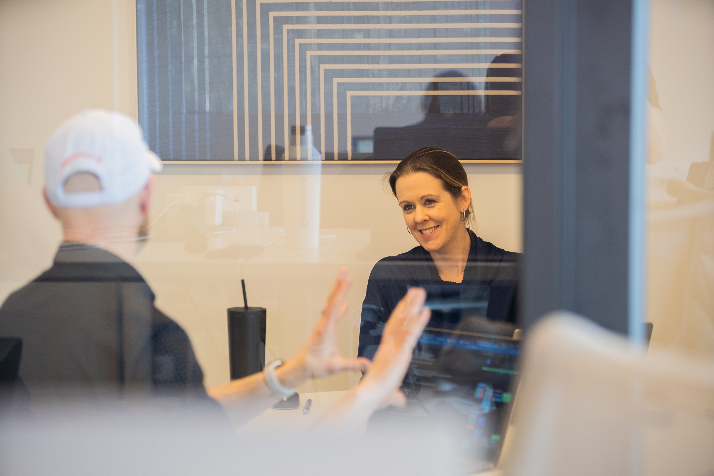 A woman smiling during a conversation with a person in a cap, viewed through a glass wall in an indoor office setting.