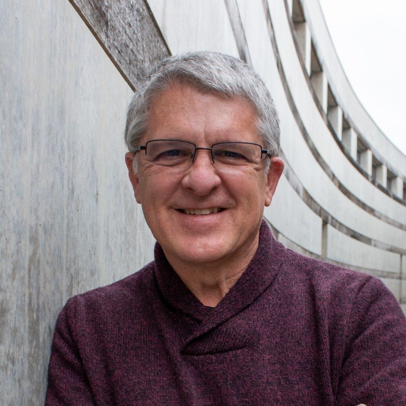 A smiling middle-aged man with gray hair and glasses standing against a curved architectural structure.