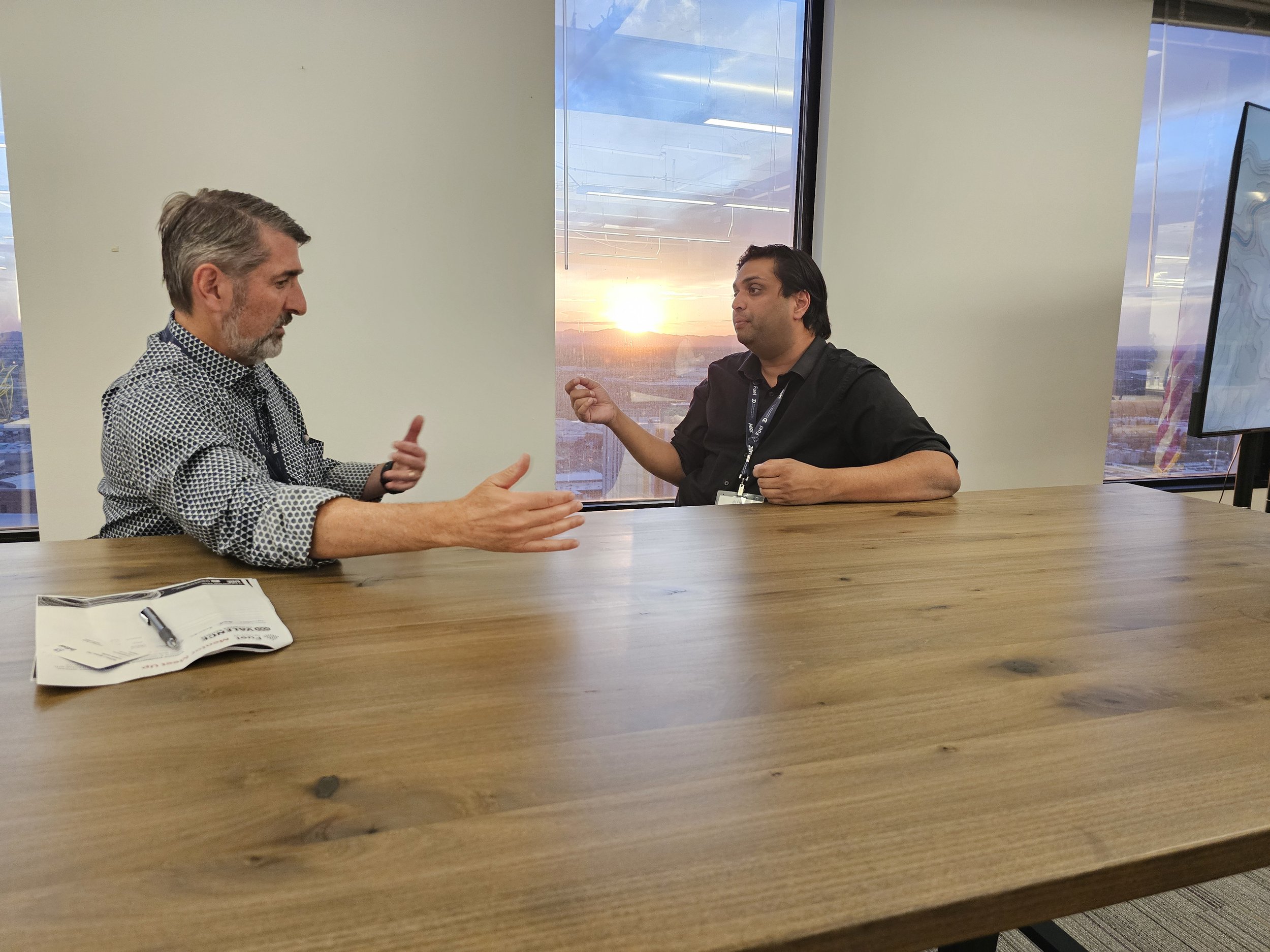 Two men having a discussion at a conference room table with a sunset view through large windows in the background.